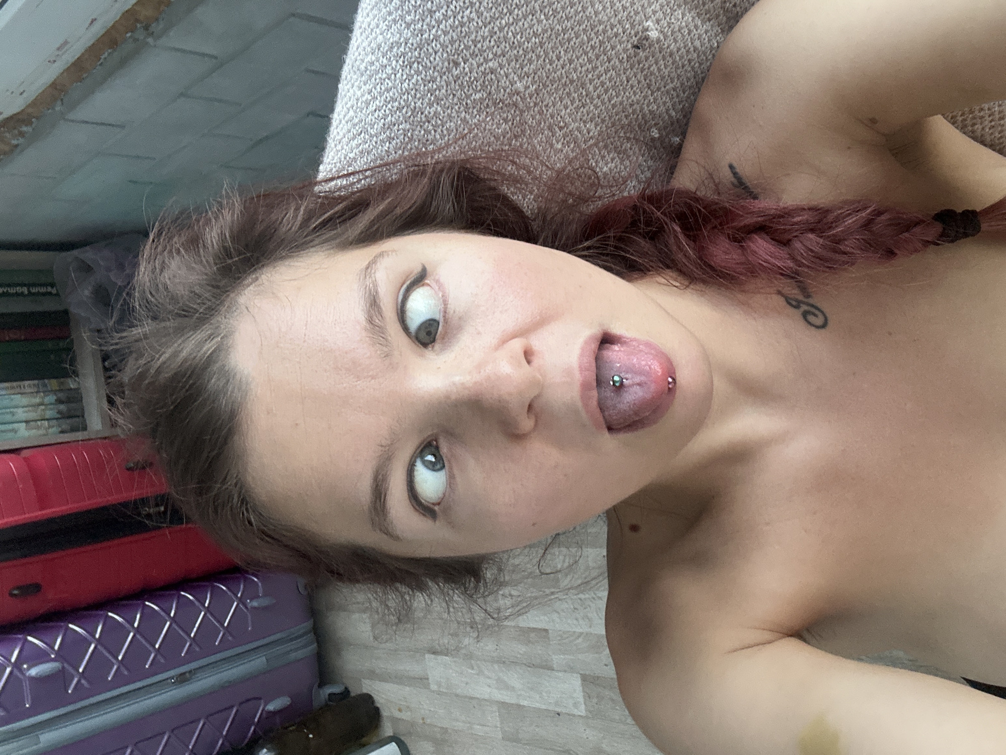 A person is lying on a light-colored couch in what appears to be an indoor setting. The person has light skin and is partially visible from the shoulders up. They have medium-length brown hair, with a portion braided and dyed a reddish color. The person's tongue is extended and has a visible piercing. The person is looking upwards, with exaggerated wide eyes. There are tattoos visible on the upper chest and near the shoulder. Adjacent to the couch, there is a stack of suitcases, red and purple. Also, a stack of books is partially visible. The flooring is light colored and appears to be a light wood or laminate. The environment gives the impression of a residential space. Vladimir, Russia is not identifiable in the picture.