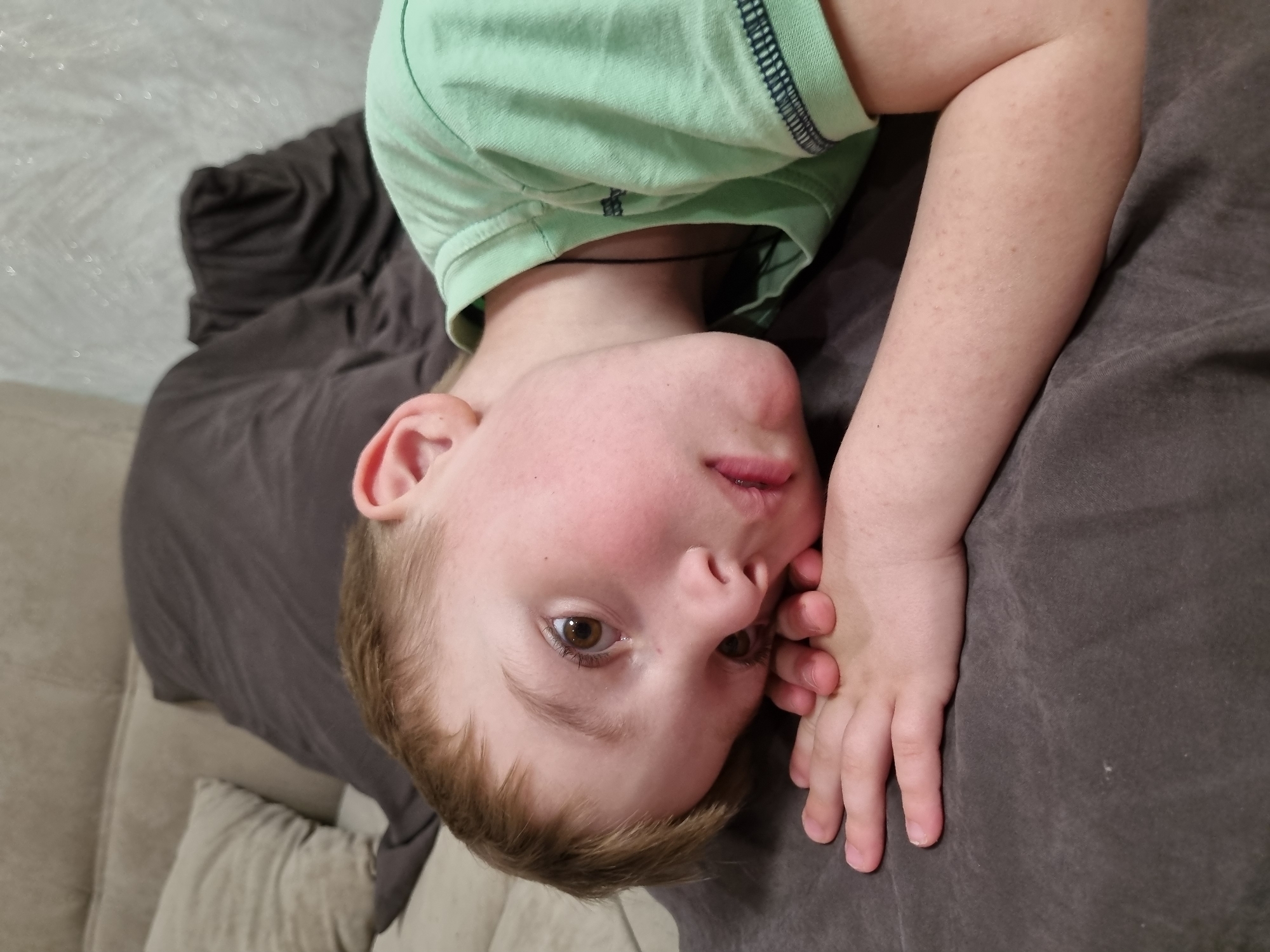 A young, fair-skinned boy is lying on his back on a dark gray fabric-covered surface, likely a couch or bed. He is wearing a light green t-shirt with dark trim around the collar and sleeves. His head is tilted upside down towards the viewer. His hair is light brown, and his eyes are brown. His right hand is near his face, resting on the gray fabric. Behind him, to the left, is a gray blanket and a portion of a light-colored couch or bed. The location is unknown and cannot be identified as Chaikovsky, Russia from the image provided.