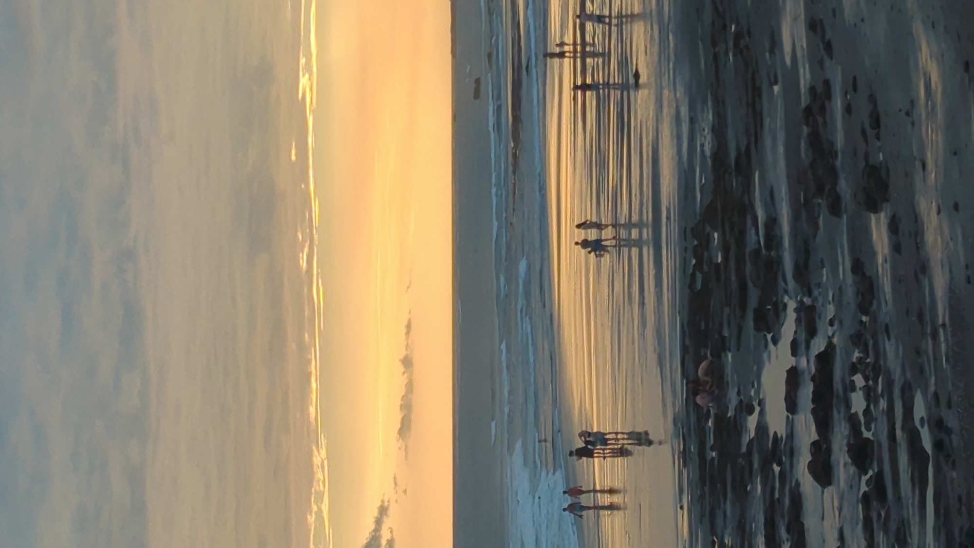 The image depicts a beach scene at sunset or sunrise, with the sun casting a warm, golden light across the sky and reflecting on the wet sand. Several people are visible walking along the shoreline, casting long shadows. The water is shallow, creating a mirror-like surface that exaggerates the elongated reflections of the figures.
The foreground of the image is dominated by dark, wet sand with pools of water and some rocks or debris. The midground shows the people walking on the beach, interspersed with the shimmering reflections of the sky and the sun. The background features the vast expanse of the sky, painted in hues of orange, yellow, and pale blue, with some wispy clouds. The overall atmosphere is serene and peaceful, suggesting a leisurely stroll on the beach during a beautiful time of day.
The location is identified as North Kuta, Indonesia. The time of day is clearly indicated by the low angle of the sun and the warm, saturated colors of the sky. There are no visible objects or text in the image. The activity is simply people enjoying a walk on the beach.