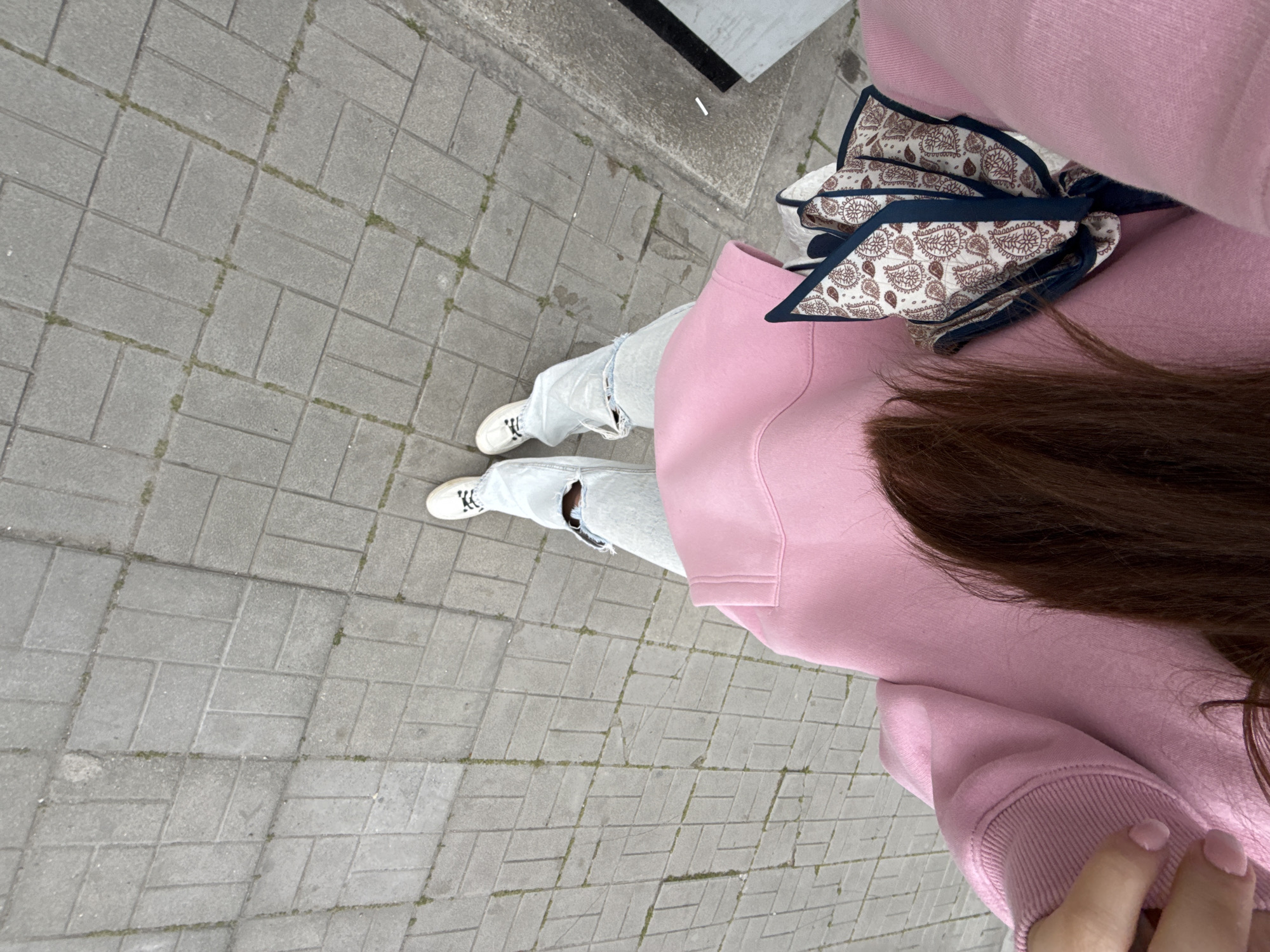 A first-person perspective shot looking down at a person standing on a paved walkway. The person is wearing a pink hooded sweatshirt, ripped light-wash jeans, and white sneakers. A paisley patterned scarf is tied around the hood. Brown hair is partially visible. The walkway is made of gray rectangular pavers. The location is not identifiable as Plovdiv, Bulgaria from visual information.
