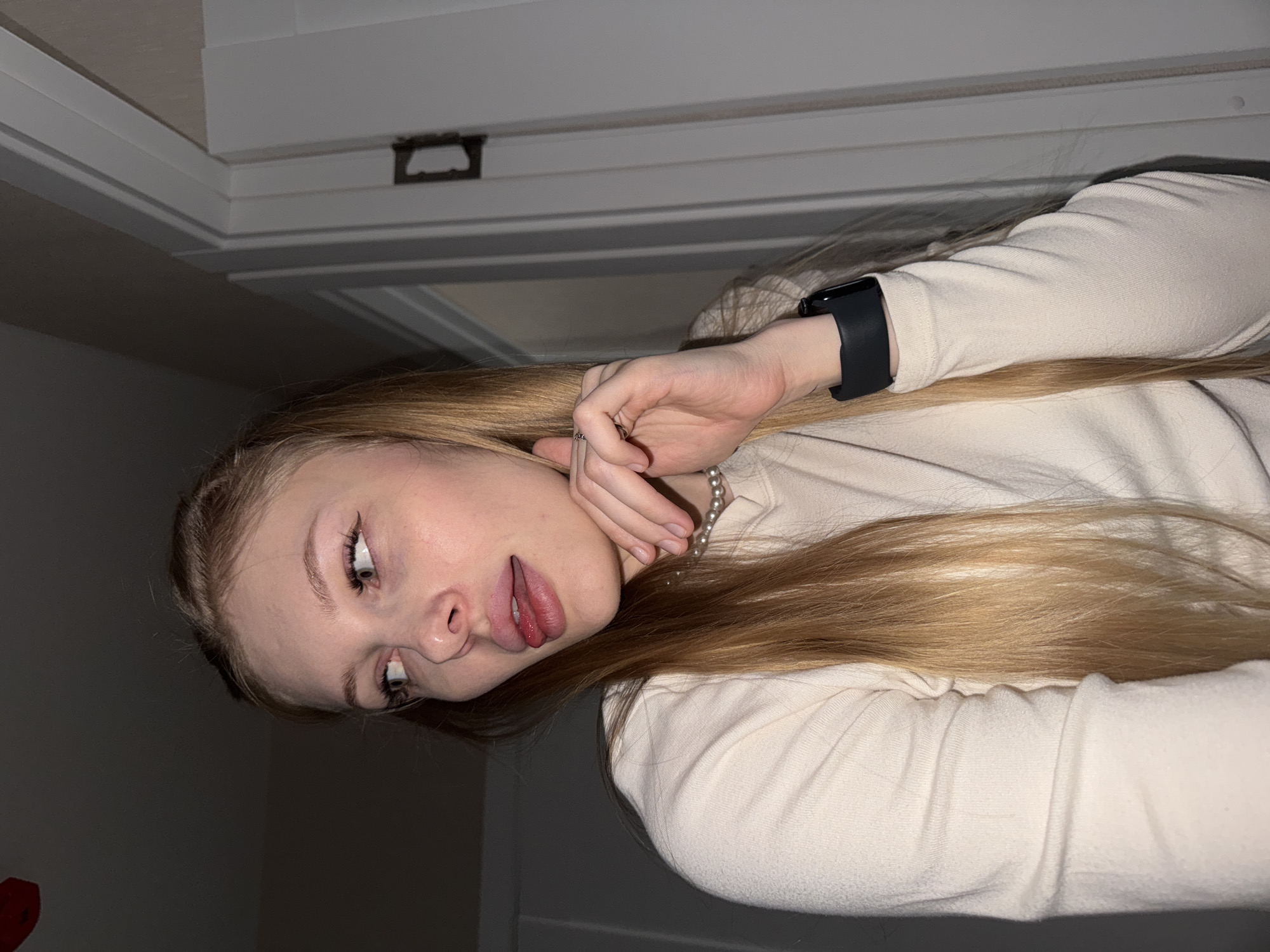 A young woman with long blonde hair is shown from the chest up, looking to her left. She is wearing a cream-colored long-sleeved shirt and a black smartwatch on her left wrist. Her right hand is raised to her face, with her fingers touching her chin and her thumb supporting her jawline. Her tongue is slightly extended. She is wearing a pearl necklace. The background is dimly lit, with a doorway and part of a wall visible on the right. The image does not provide any contextual clues that would indicate its location in the city of Perm, Russia.