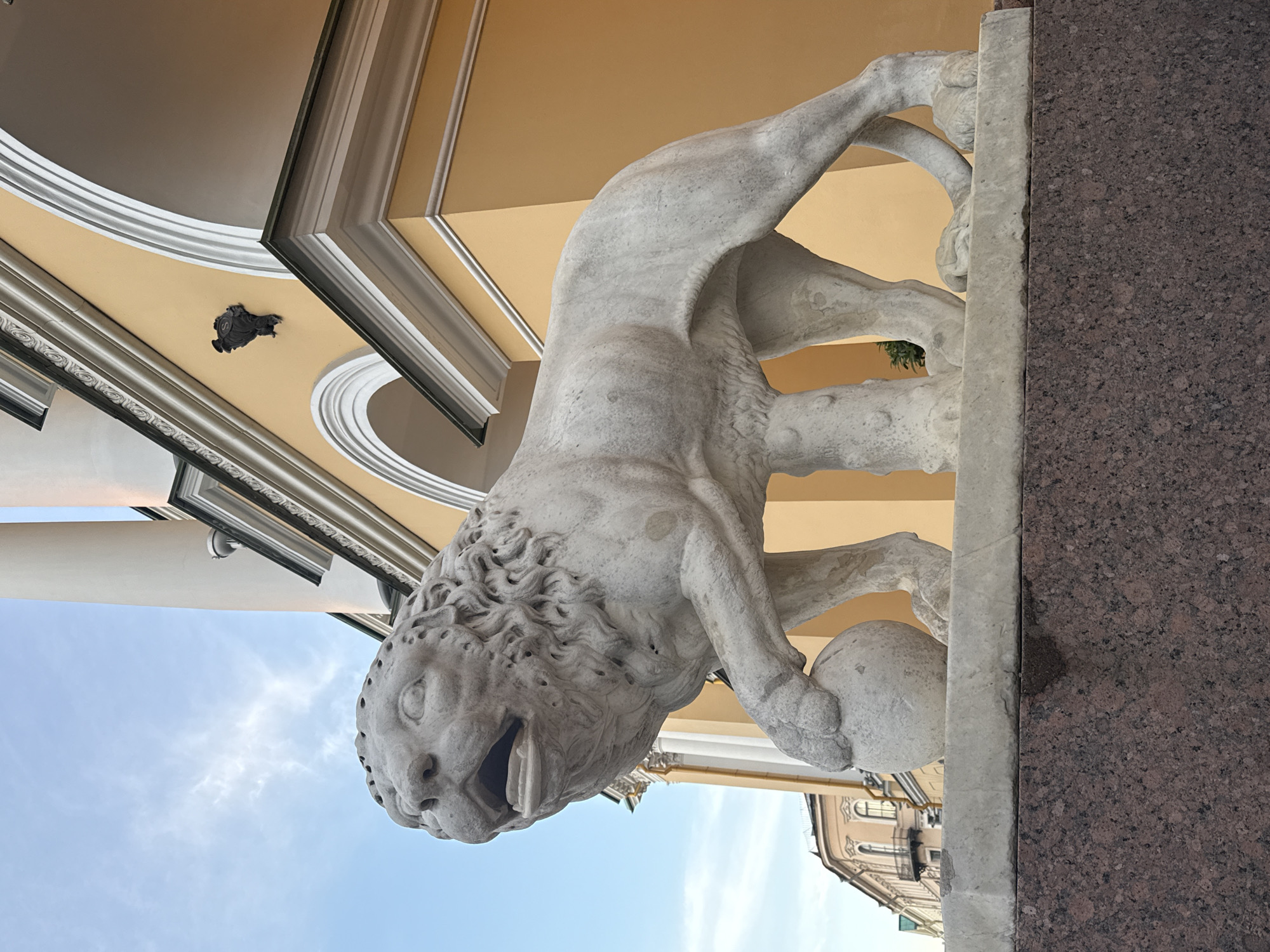An ornate stone sculpture of a lion with its paw resting on a sphere stands on a granite pedestal in front of a building with a yellow facade in Saint Petersburg, Russia. The lion sculpture is detailed, with a mane, open mouth, and visible teeth. The building behind it features large white columns and arched entrances, with decorative white trim. The sky above is blue with some wispy white clouds. The overall scene suggests an architectural or monumental setting.