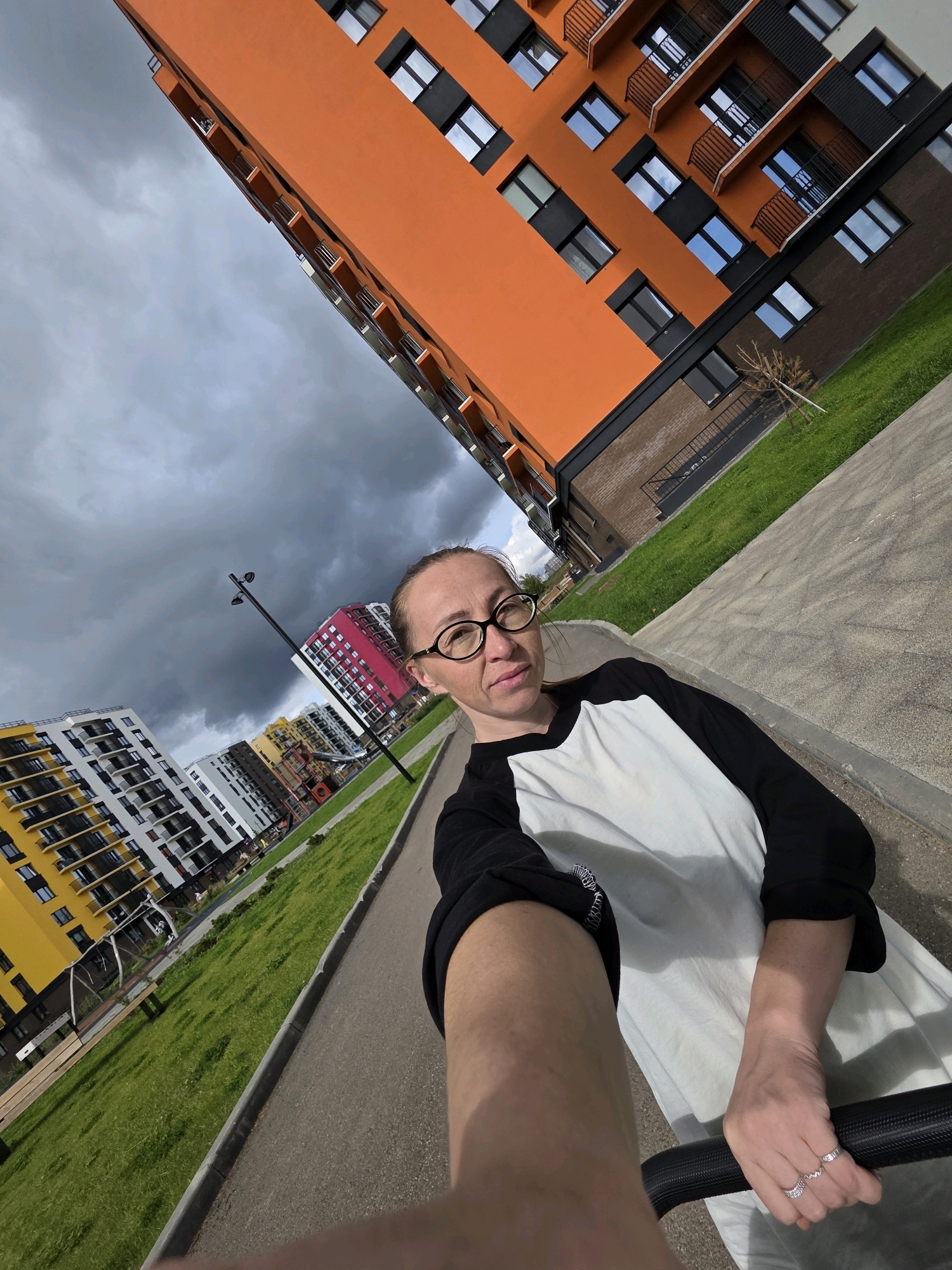In Pervomaysky, Russia, a woman with glasses and a black and white shirt is captured in a selfie outdoors, positioned on a paved pathway. She is holding onto a black object, likely a stroller, with her right hand, adorned with two rings. Behind her, a modern apartment complex with an orange facade dominates the view, its windows reflecting the cloudy sky. To the left, other residential buildings with yellow and white facades are visible, along with a grassy area and a lamppost. The overall scene depicts a residential area with contemporary architecture under an overcast sky.