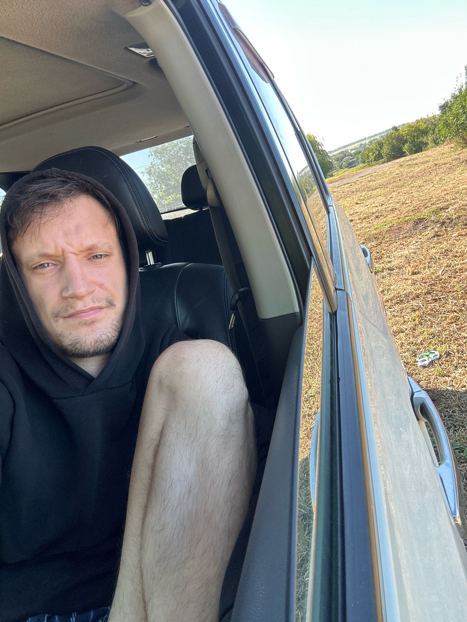 A man with light skin, brown hair, and a beard is sitting in the driver's seat of a car, looking towards the camera with a neutral expression. He is wearing a black hooded sweatshirt and his right leg is visible, resting on the passenger seat. The car's interior is visible, including the black leather seats and a sunroof. The car is parked outdoors, with a dry, grassy field and trees visible through the open passenger door. In the background, there are rolling hills and a clear sky. The scene appears to be in the city of Makiv, Ukraine, given the context.