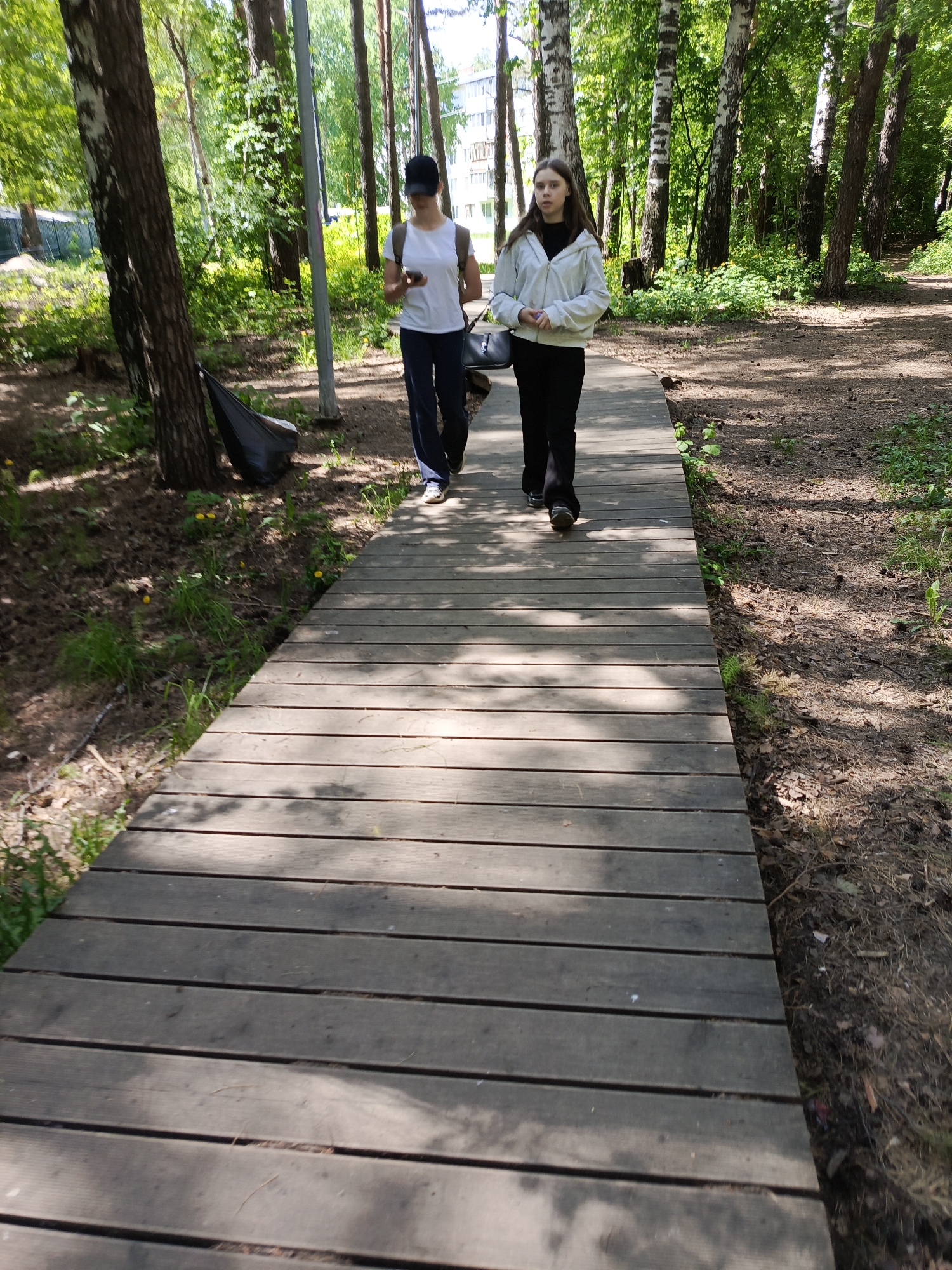 Two individuals are walking on a wooden boardwalk in a wooded area, likely in Chaikovsky, Russia. The person on the left is wearing a white t-shirt, dark pants, a black baseball cap, and appears to be looking at a mobile device. They are also carrying a dark-colored bag. The individual on the right is wearing a white jacket and dark pants. The boardwalk is constructed of wooden planks and runs through a forest or park-like setting. The surrounding environment is characterized by trees and ground covered with pine needles and grass. The lighting suggests it is daytime.