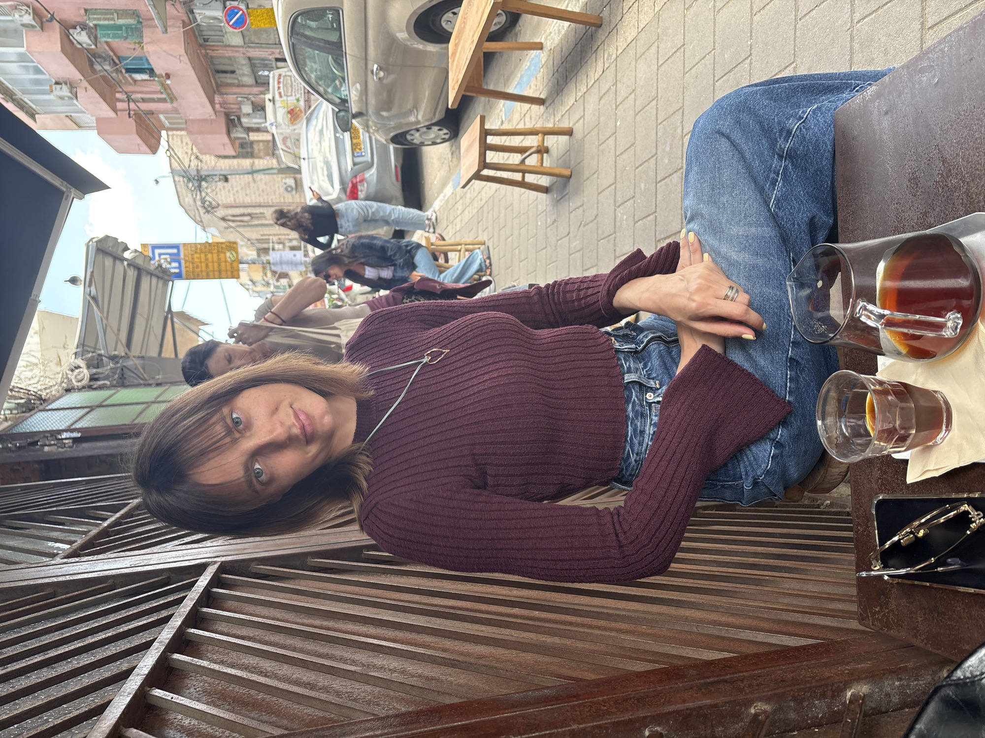 A woman wearing a maroon sweater and jeans sits at an outdoor cafe, visible from the waist up. She is positioned diagonally in the frame, with her head tilted slightly toward the viewer. A pitcher and a glass of dark liquid, possibly tea or coffee, sit on the table beside her. A pair of glasses in a case is also on the table. In the background, other people are seated at tables along the street, and a car is parked nearby. Buildings line the street, suggesting an urban setting. The location is Tel Aviv, Israel.