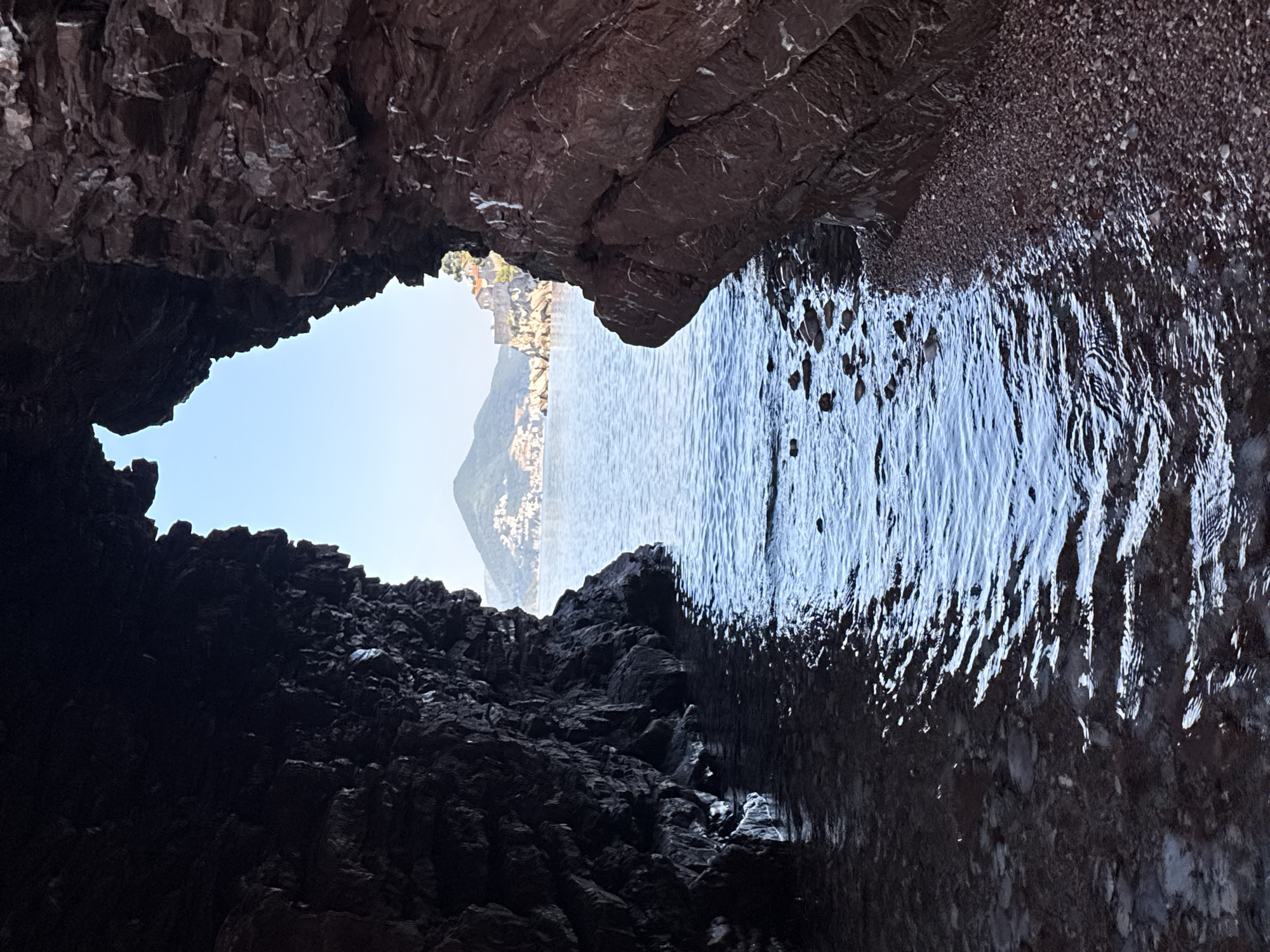 The image captures a view from within a rock formation, framing the sea and a distant hillside town. The foreground is dominated by the dark, textured interior of the rock, with water lapping at the base of the formation on the left. The right side of the foreground consists of a pebbled shore. Beyond the opening in the rocks, the sea stretches out, calm with gentle ripples. In the distance, across the water, a hillside is covered with buildings, characteristic of Sveti Stefan, Montenegro. A prominent peak rises behind the town. The sky is clear and pale blue. There are no people or animals visible in the scene.