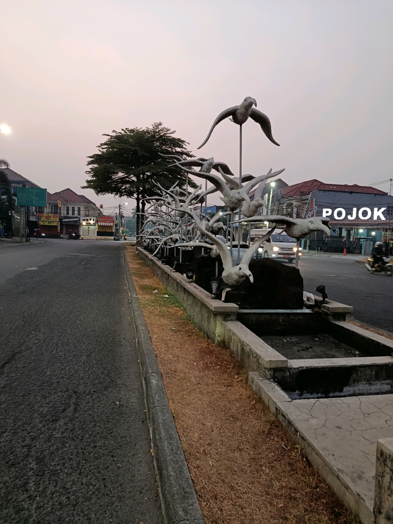 The image shows a public art installation of bird sculptures in a street in the city of Bekasi, Indonesia. The sculptures are made of a light-colored material and depict birds in flight. They are arranged in a row along a concrete planter box filled with dry grass and what appears to be a dried-up water feature.
In the background, there is a street with asphalt paving, a sidewalk, and several buildings. The buildings have a mix of architectural styles, with some appearing to be shops or commercial establishments. A tree with dense foliage stands behind the bird sculptures. The sky is overcast with a soft, diffused light, suggesting it is either early morning or late evening.
There are a few vehicles visible on the street, including a white van and a motorcycle. A person is riding the motorcycle. The word "POJOK" is prominently displayed in large white letters on one of the buildings in the background. This likely indicates a location or business name.
The overall impression is a street scene in a moderately developed urban area, with a focus on a distinctive piece of public art. The environment appears to be relatively calm, with little pedestrian activity.