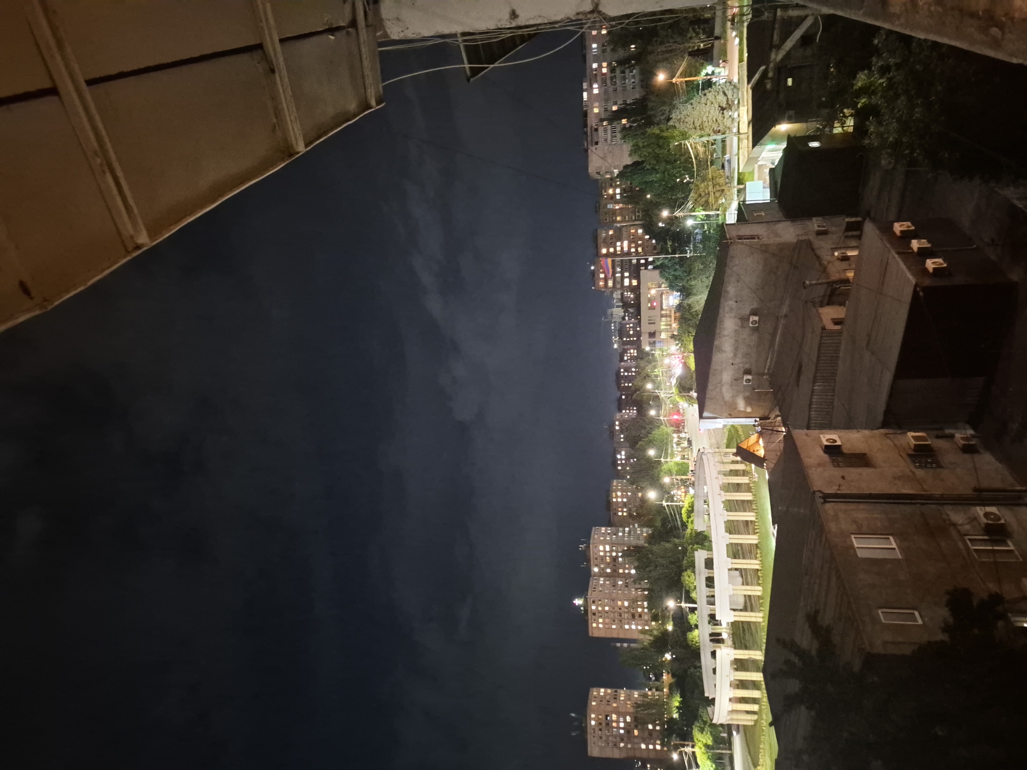 The image is a nighttime view from a balcony, looking out over a cityscape. The foreground features the edge of a building's roof and a dark, textured surface. Beyond this, a sprawling urban scene unfolds, characterized by numerous apartment buildings with illuminated windows. Trees and streetlights are interspersed throughout the urban landscape, suggesting a residential area with public spaces. The sky is dark and cloudy.
The activity depicted is the general life of a city at night, with residential buildings lit up, indicating people are indoors. The time of day is clearly night, evidenced by the dark sky and the artificial lighting from buildings and streetlights. The weather appears to be clear enough for an urban view, despite the cloudy sky, as visibility is good. Location cues suggest an urban setting, likely a densely populated area with multi-story apartment blocks. There are no visible people or specific events taking place. The overall mood is calm and observational, capturing the quiet ambiance of a city after dark. No visible text is present in the image. The context provided indicates the location is Yerevan, Armenia.