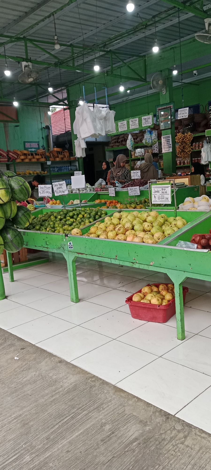 An outdoor fruit stand in Maguwoharjo, Indonesia, features a variety of produce displayed on green shelves. Watermelons are stacked on the left, with oranges, limes, and guavas filling the central shelves. In the background, three women in hijabs are attending to customers. The stand is covered by a metal roof, and the floor is tiled. A sign indicates a toilet is available.