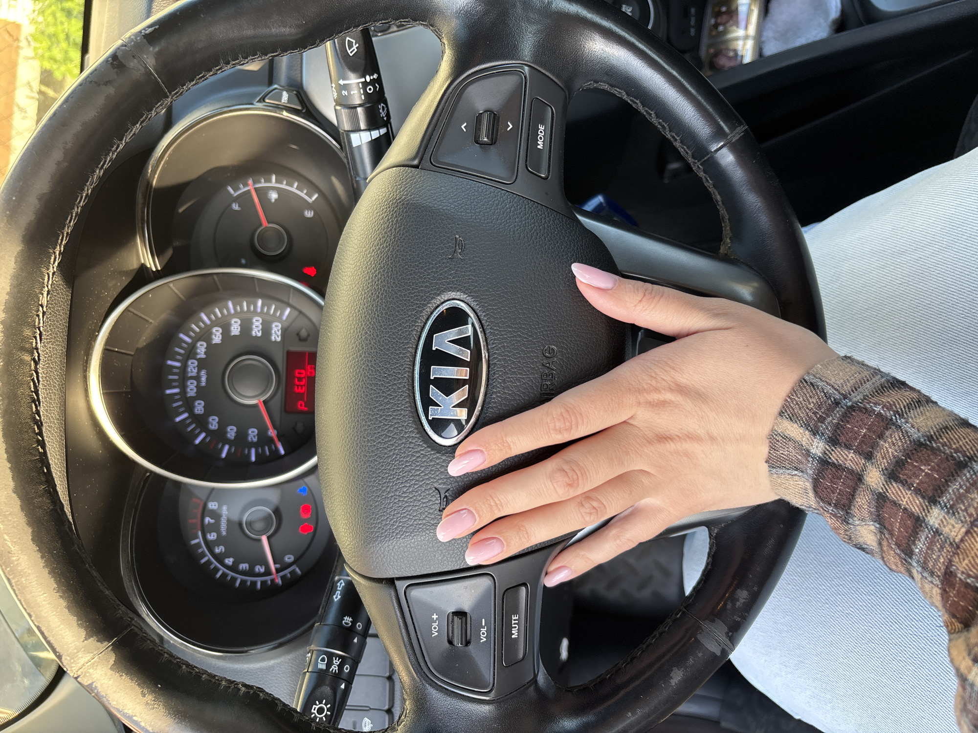 A close-up shot from the perspective of a driver shows a person's right hand resting on the steering wheel of a Kia car. The hand has manicured nails with light pink polish and is partially covered by the sleeve of a brown and tan plaid shirt, with light blue jeans visible below. The black leather steering wheel appears somewhat worn, especially on the top left.
The car's dashboard is visible behind the steering wheel, featuring a speedometer marked up to 220 km/h and a tachometer up to 8 x1000rpm. Several dashboard lights are illuminated, including a red fuel warning light indicating low fuel, a blue temperature light, and another red warning indicator. The text "P_ECO" is also illuminated in red on the dashboard. Integrated controls on the steering wheel include buttons for "MODE", "VOL+", "VOL-", and "MUTE", as well as navigation buttons. The central horn pad features the "KIA" logo and partially visible "AIRBAG" text. Wipers and light stalks with various symbols are also visible. The bright lighting suggests it is daytime. The setting is inside a car in Rusanovo, Russia.