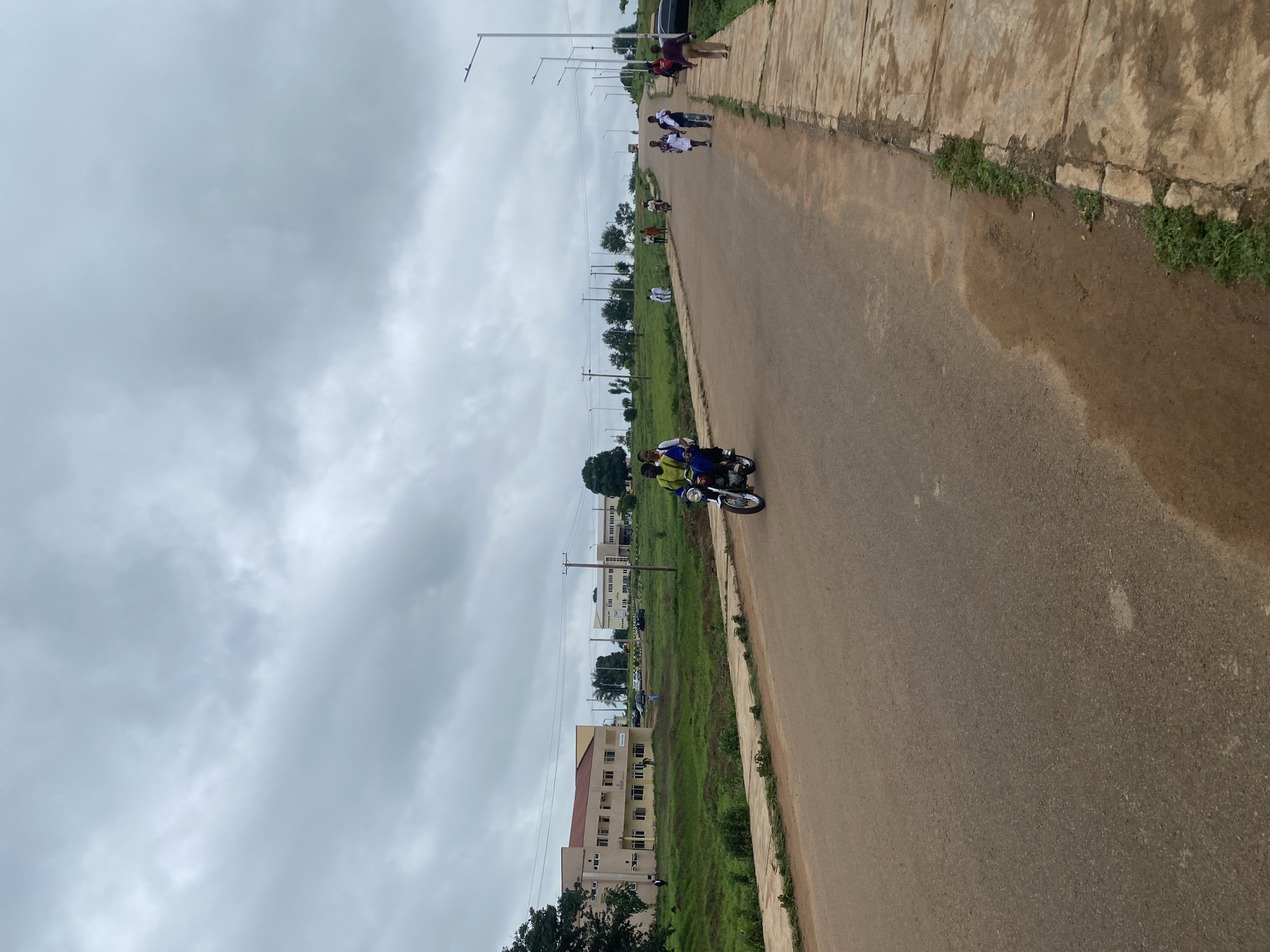 In Lafia, Nigeria, under a cloudy sky, an asphalt road runs through a landscape dotted with buildings and lush green grass. On the road, a motorcycle carrying two individuals is moving towards the viewer. To the right of the road, a concrete sidewalk stretches into the distance, lined with streetlights. Several pedestrians are walking on the sidewalk, some moving away from the viewer. In the background, low-rise buildings are visible amidst trees and grassy areas. The overall scene suggests a developing urban or institutional environment.