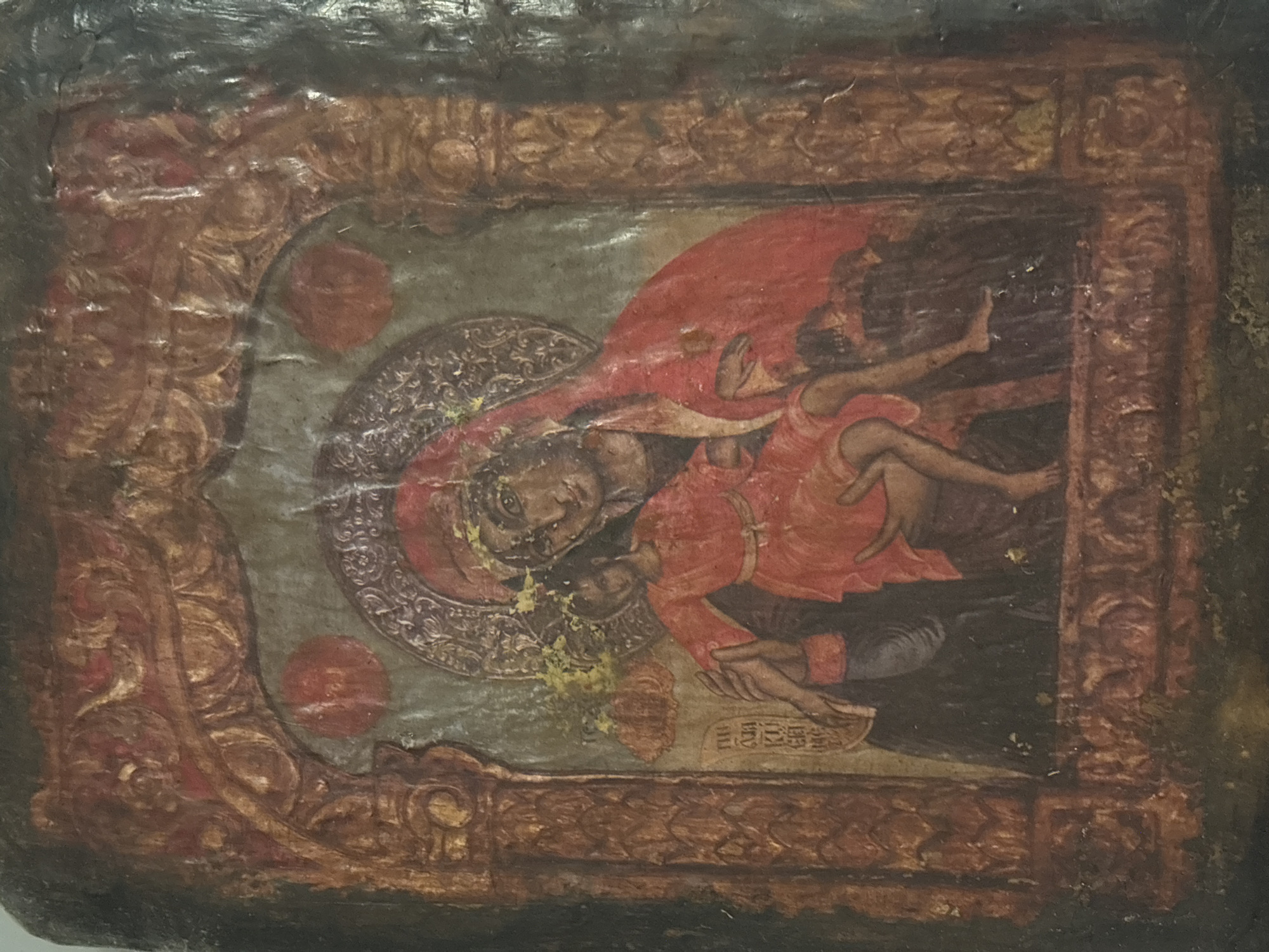 The image displays an aged religious icon, characteristic of Orthodox Christian iconography, likely depicting the Virgin Mary holding the infant Jesus. The painting is encased within an elaborate, dark red and gold-toned decorative border, suggesting a heavily ornamented frame or riza. The style, consistent with Byzantine or Post-Byzantine art, is commonly found in regions such as Sofia, Bulgaria.
The two central figures are rendered with stylized features and positioned diagonally. The Virgin Mary, on the left, gazes outwards with a serene, contemplative expression. A detailed, patterned halo-like disc, adorned with geometric designs, encircles her head. She is draped in a flowing red mantle (maphorion) over a darker garment. The Christ child, dressed in a red tunic, is held in her arms, depicted in a dynamic pose with arms and legs outstretched, indicating movement. A faint halo is also visible around the child's head.
The background of the central image is a muted greenish-gray. The icon shows significant signs of age and wear, including visible cracks, discoloration, flaking paint, and areas of paint loss, particularly around the Virgin's face and the lower center. Faint, stylized inscriptions, appearing to be Cyrillic script, are discernible below the child's feet within the main image area. The ornate border features intricate swirling and geometric patterns in shades of gold, brown, and red, enhancing the icon's reverential quality. Several indistinct red circular elements are also integrated into the border design.