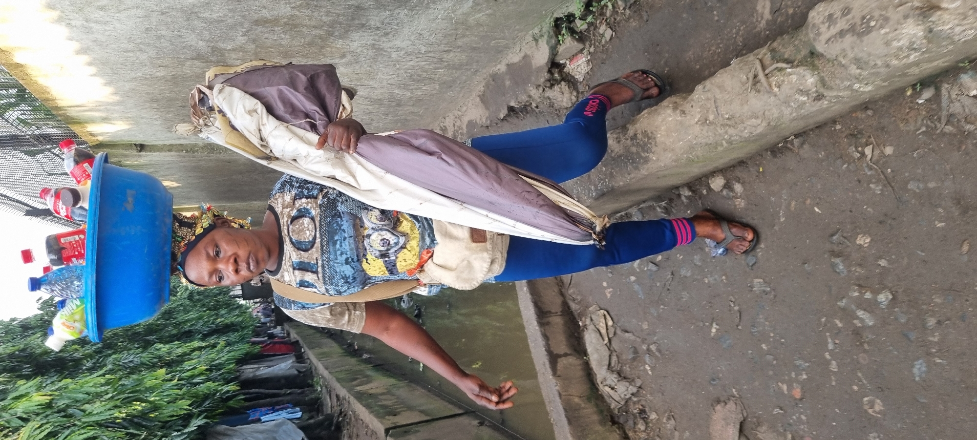 In Ikeja, Nigeria, a woman walks along a muddy path beside a concrete drainage ditch, balancing a large blue tub filled with assorted bottled drinks on her head. She wears a patterned t-shirt, blue leggings with pink stripes at the ankles, and flip-flops. A light-colored crossbody bag is slung over her shoulder, and she carries a rolled-up umbrella in her right arm. The environment is outdoor, with a concrete wall to her right and lush green foliage and hanging clothes to her left. The ground is dirt and debris-strewn, and the overall scene suggests an urban, potentially informal market setting.