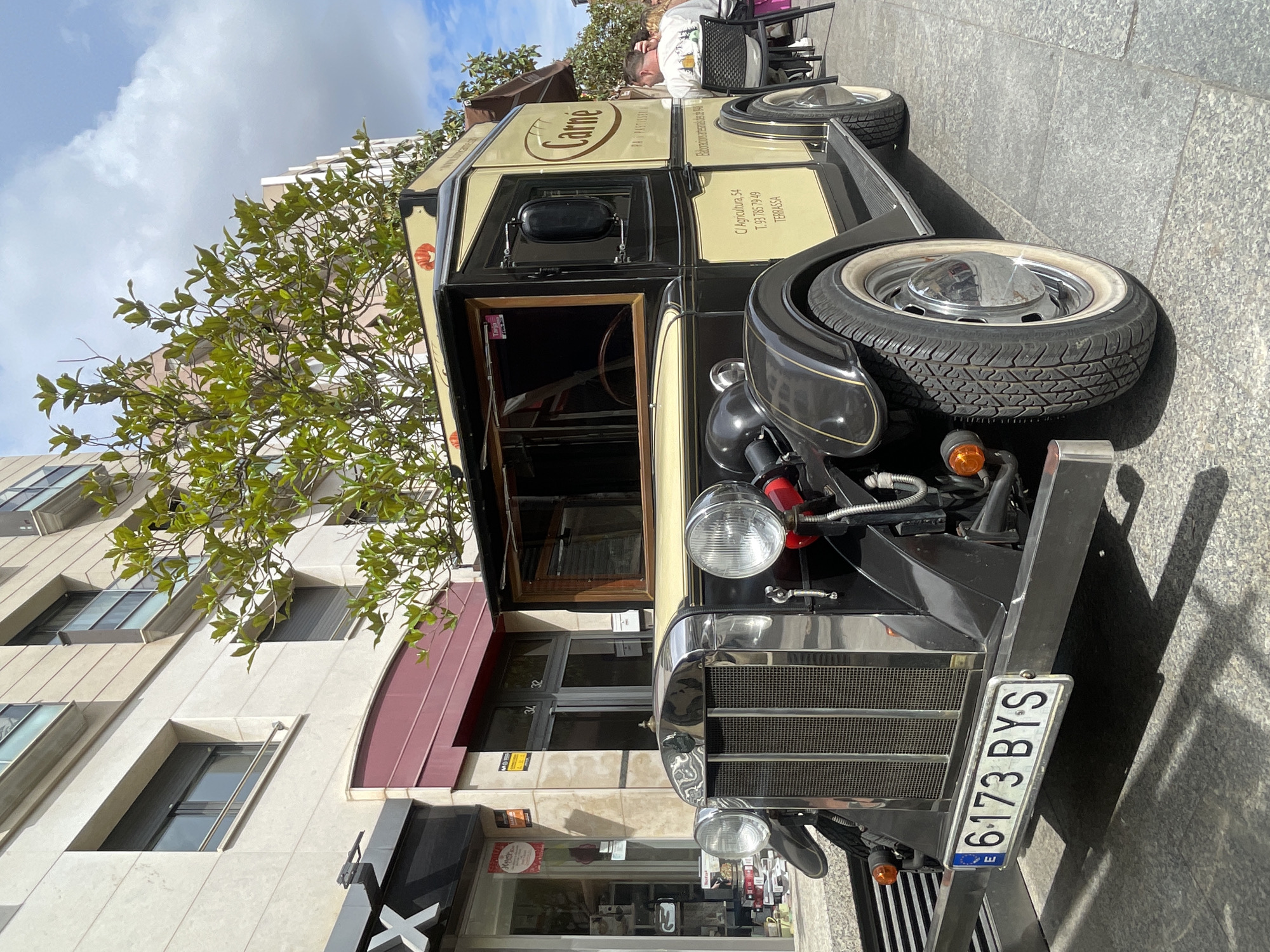 The image shows a vintage vehicle parked on a street in Terrassa, Spain. The vehicle has a black and cream color scheme and features the word "Carne" written on its side. The license plate number is "6173 BYS". A tree is partially obscuring the vehicle, and a multi-story building is visible in the background. A sign with "C/ Agricultura, 54 T.93 785 7949 TERRASSA" is also visible on the vehicle. A person is seen sitting at a table nearby. A shop with a logo, partially marked with an "X," is also located in front of the building.