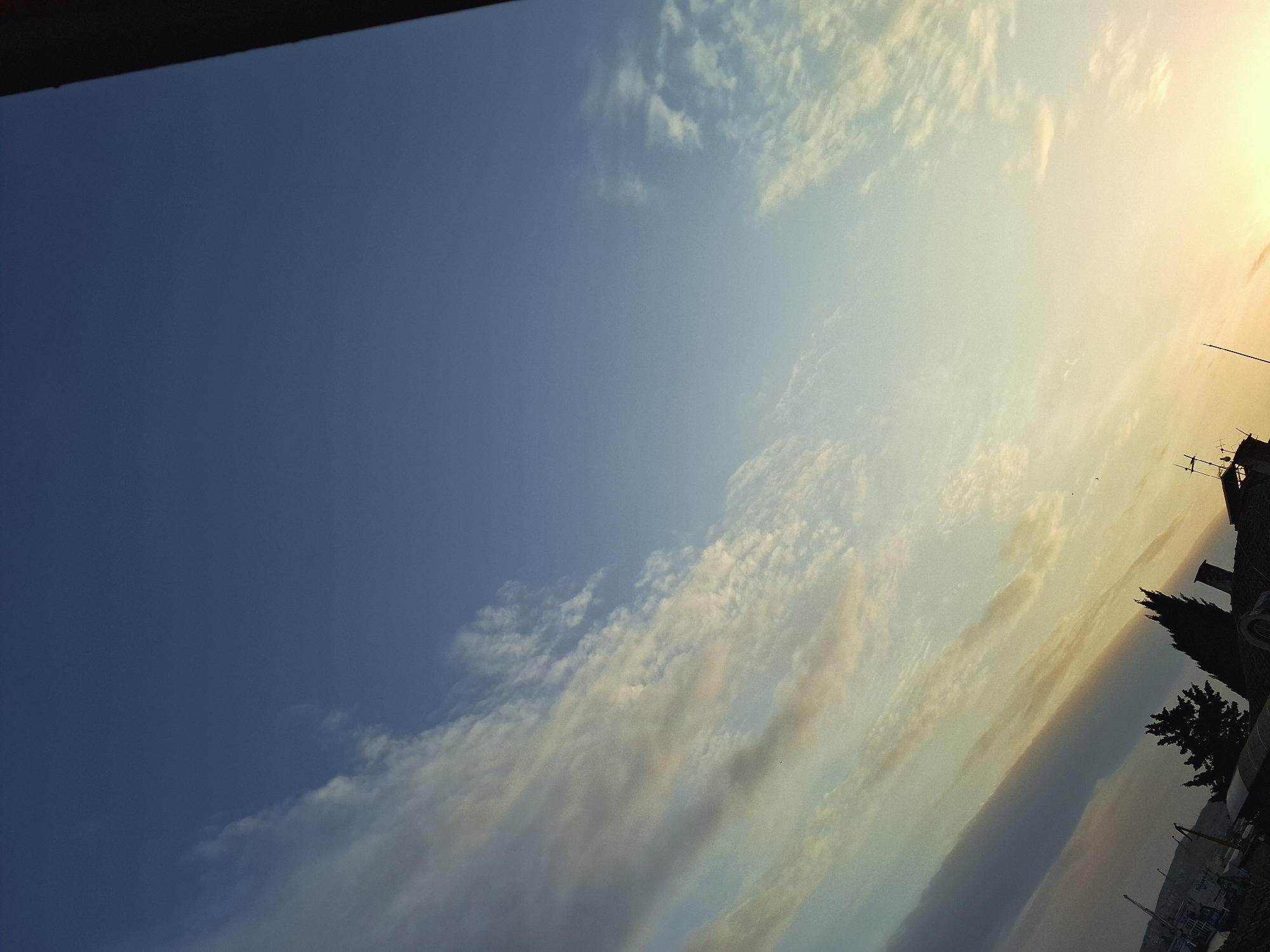 The image shows a view of the sky with a building, crane and tree in the bottom right corner of the frame. The sky features different shades of blue, from dark to light, and some clouds. There is a sunset glow visible near the building in the bottom right corner. The image is rotated at an angle. It is not possible to confirm the location of the city.