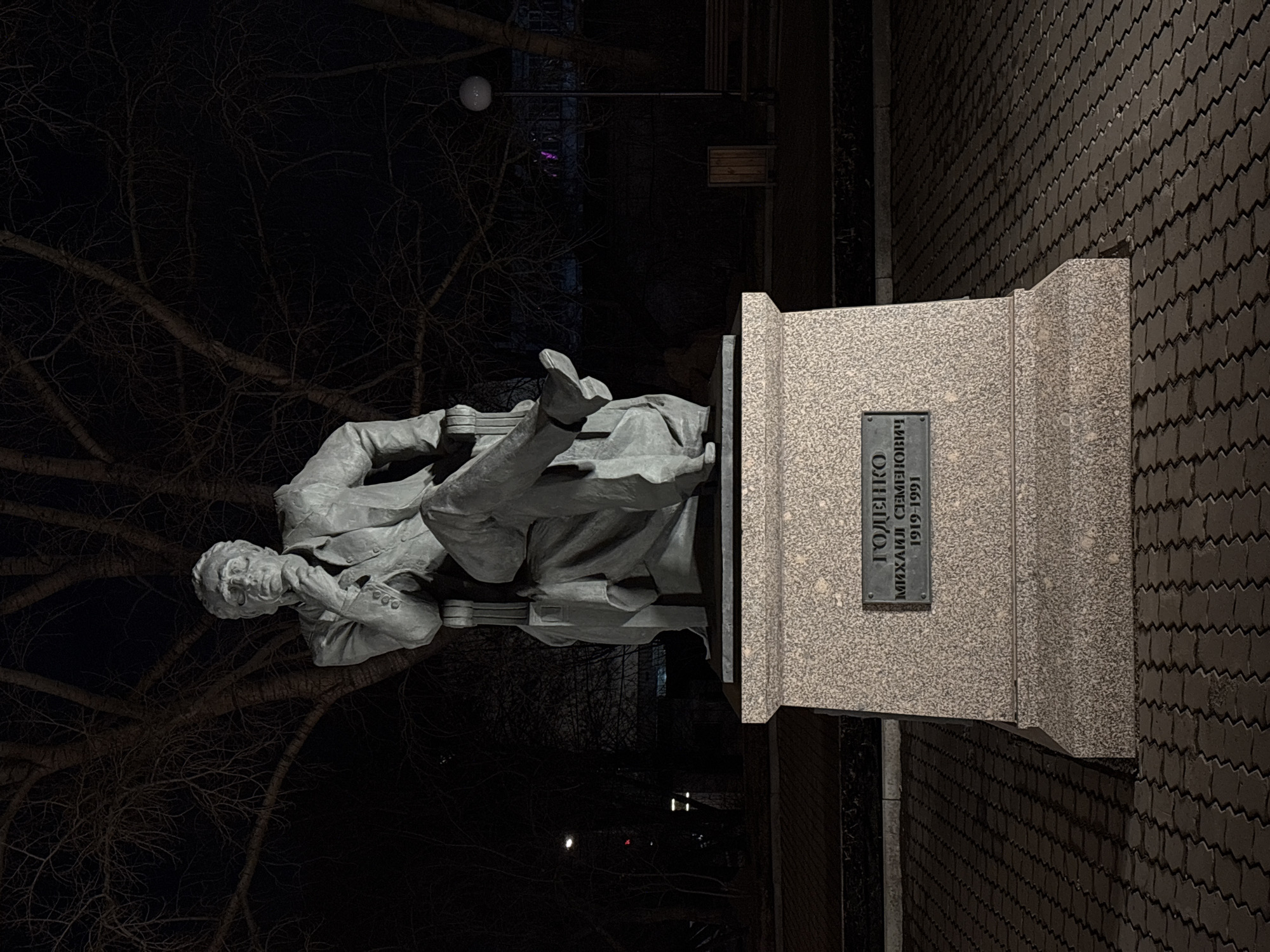 The image captures the monument to Mikhail Semenovich Godenko, a Russian choreographer, located in Krasnoyarsk, Russia. The monument consists of a sculpture of Godenko in a seated position, attached to a stone pedestal. The pedestal bears a plaque with his name and dates, "Годенко Михаил Семенович 1919-1991". The statue and pedestal are made of a light-colored stone. The scene is dimly lit, likely at night. In the background, bare branches of trees are visible. The area surrounding the monument is paved with dark-colored bricks. The image is oriented sideways, with the monument appearing to be rotated 90 degrees clockwise.
