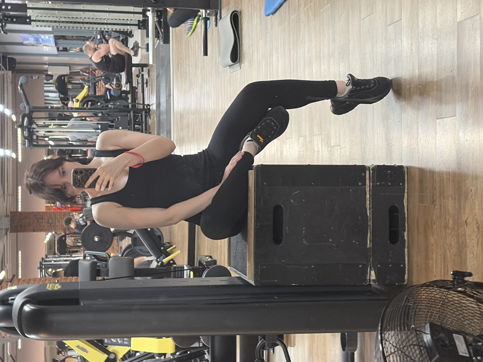 A young woman with dark hair is taking a mirror selfie in a gym. She is wearing a black tank top and black leggings, and black sneakers with yellow accents. She is sitting on a black plyometric box, with her right leg extended and her left leg bent at the knee. Her left hand is resting on her left thigh, and her right hand is holding a smartphone, capturing the reflection. In the background, other gym equipment and people can be seen. The gym has wooden flooring and exposed brick walls, suggesting a modern industrial design. The overall lighting is bright, typical of a gym environment. The image is set in a gym located in the city of Chaikovsky, Russia.