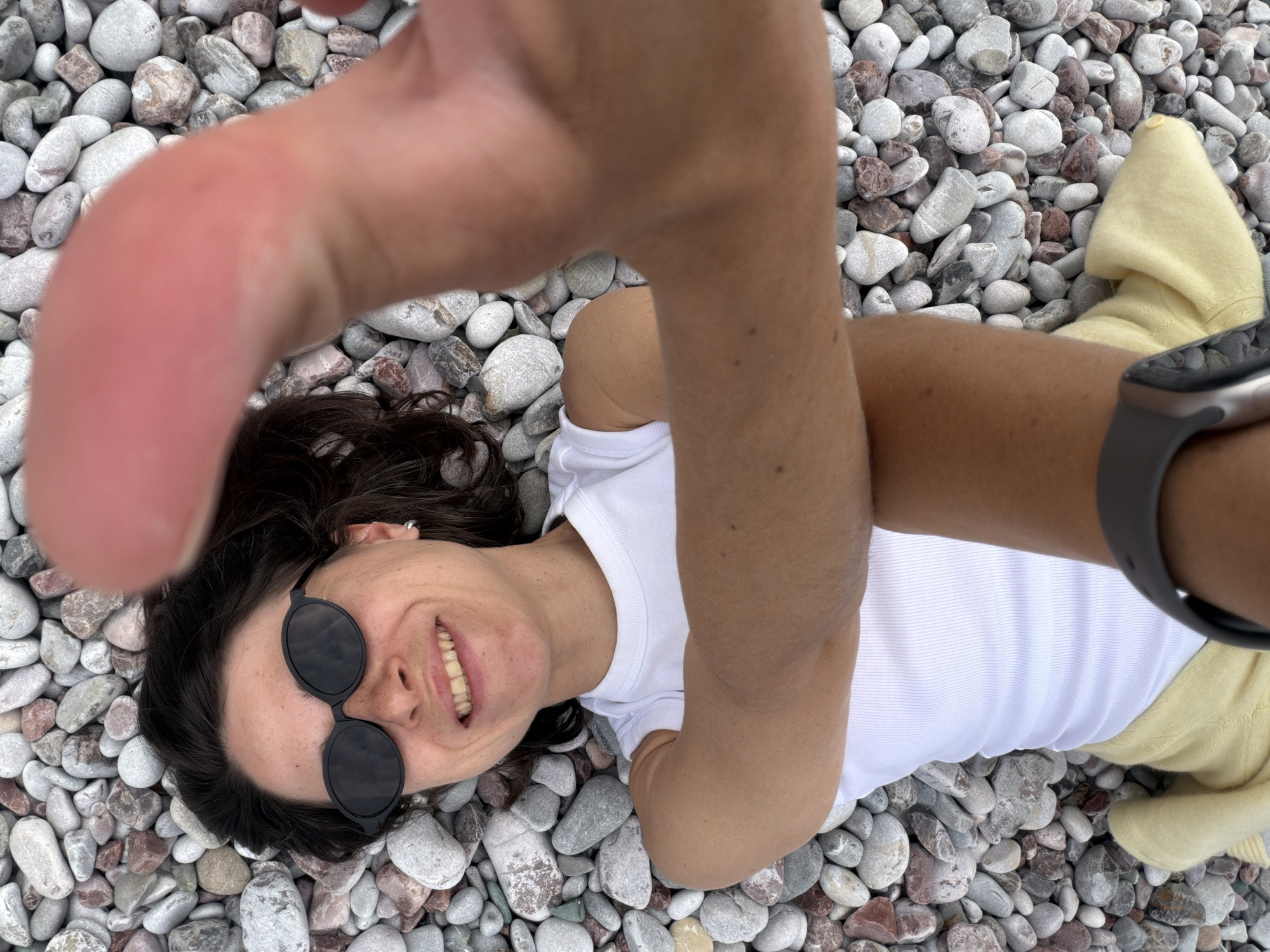 The image shows a woman lying on a pebble beach in Rafailovići, Montenegro. She is wearing a white shirt, black sunglasses, and a gray smartwatch. Her hair is dark and curly. She is smiling at the camera, which is held at arm's length, with a hand partially obscuring the view. A yellow sweater is visible on the right side of the frame. The ground is covered in small, rounded pebbles of various colors.