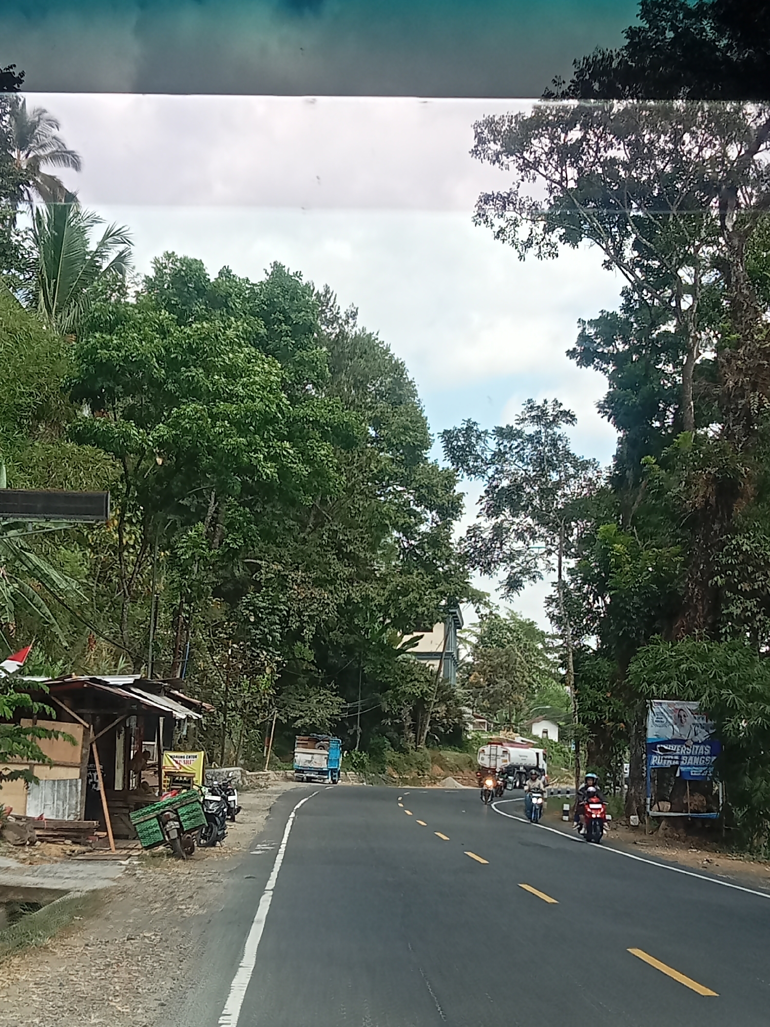 The image depicts a scenic road in Gunungtawang, Indonesia, bordered by lush green trees. The road is paved with asphalt and marked with yellow lines. Several vehicles are visible on the road, including a blue truck in the distance, three motorcycles with riders, and a white tanker truck. On the side of the road, there is a small roadside stall with wooden structures and some wooden planks displayed. A few motorcycles are parked near the stall. The sky is partly cloudy, with hints of blue sky visible. The overall scene suggests a rural or semi-rural setting with active local transportation.