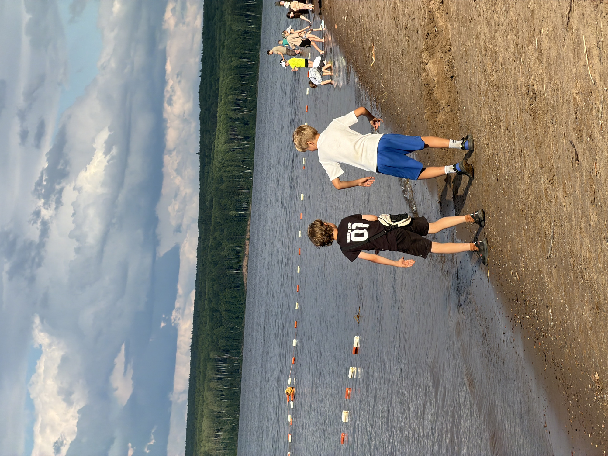 This media file depicts a group of young people at the edge of a body of water, likely a lake or a large river, in Chaikovsky, Russia. The scene is set on a sandy shoreline with a dense forest bordering the water in the background. The sky is filled with dramatic, layered clouds, suggesting a dynamic weather system, possibly with sunlight breaking through.
Several individuals are visible, mostly children and teenagers, engaged in various activities. Some are wading in the shallow water, while others are standing on the bank. Two boys are prominently featured in the foreground. One is wearing a white t-shirt and blue shorts, looking out towards the water. The other, in the foreground and slightly closer to the camera, is wearing a black t-shirt with white lettering and black shorts, also facing away from the viewer. A line of buoys stretches across the water, possibly marking a swimming area or a boundary.
The overall mood appears to be relaxed and recreational, consistent with a day spent outdoors at the water's edge. The lighting suggests it is late afternoon or early evening, given the angle of the light and the potential for long shadows, though shadows are not clearly visible in this orientation. The environment, with the forest and the calm water, points to a natural, possibly rural or suburban, setting. The text "NUMBER ONE" is visible on the black t-shirt of one of the boys in the foreground.