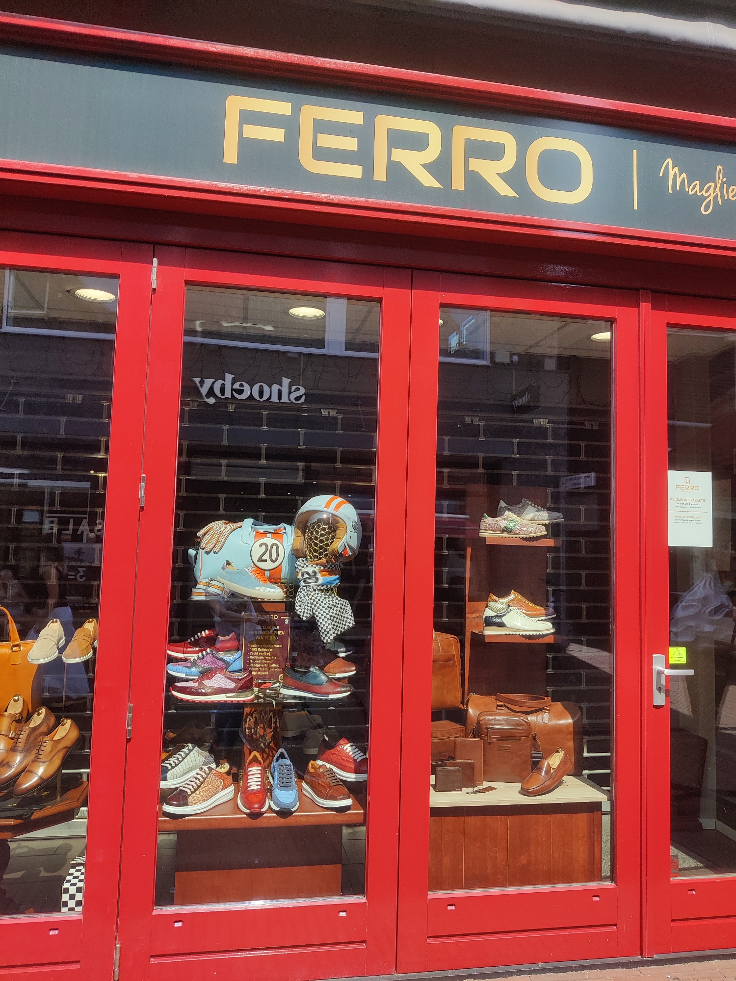 The image shows the exterior of a retail store with red double doors and large glass windows. Above the doors, a sign with gold lettering reads "FERRO | Maglie". The storefront is displaying various items, including shoes, handbags, and accessories, in the windows.
In the left window, several pairs of men's dress shoes are displayed on a dark wooden shelf. To the right of these shoes, a bright orange handbag is visible. In the center of the window, a mannequin head wearing a racing helmet with a "20" on it is displayed, along with a patterned scarf. Below this, rows of colorful athletic shoes are arranged on shelves. A black and white checkered pattern is visible on the floor of the display.
In the right window, several leather handbags and wallets are displayed on wooden shelves. A pair of women's shoes with a white sole and tan leather upper is also visible. Above these, a sign for "FERRO" with the text "WIJ ZIJN MET VAKANTIE" (We are on vacation) is displayed, indicating the store's closure.
The background of the store windows appears to be a dark brick wall. The overall scene suggests a shoe and clothing store with a focus on fashion and accessories.
Specific to Winterswijk, Netherlands, this storefront is located at Vredenseweg 118, 147, 149, where the store "Ferro Schoenen & Mode" is situated.