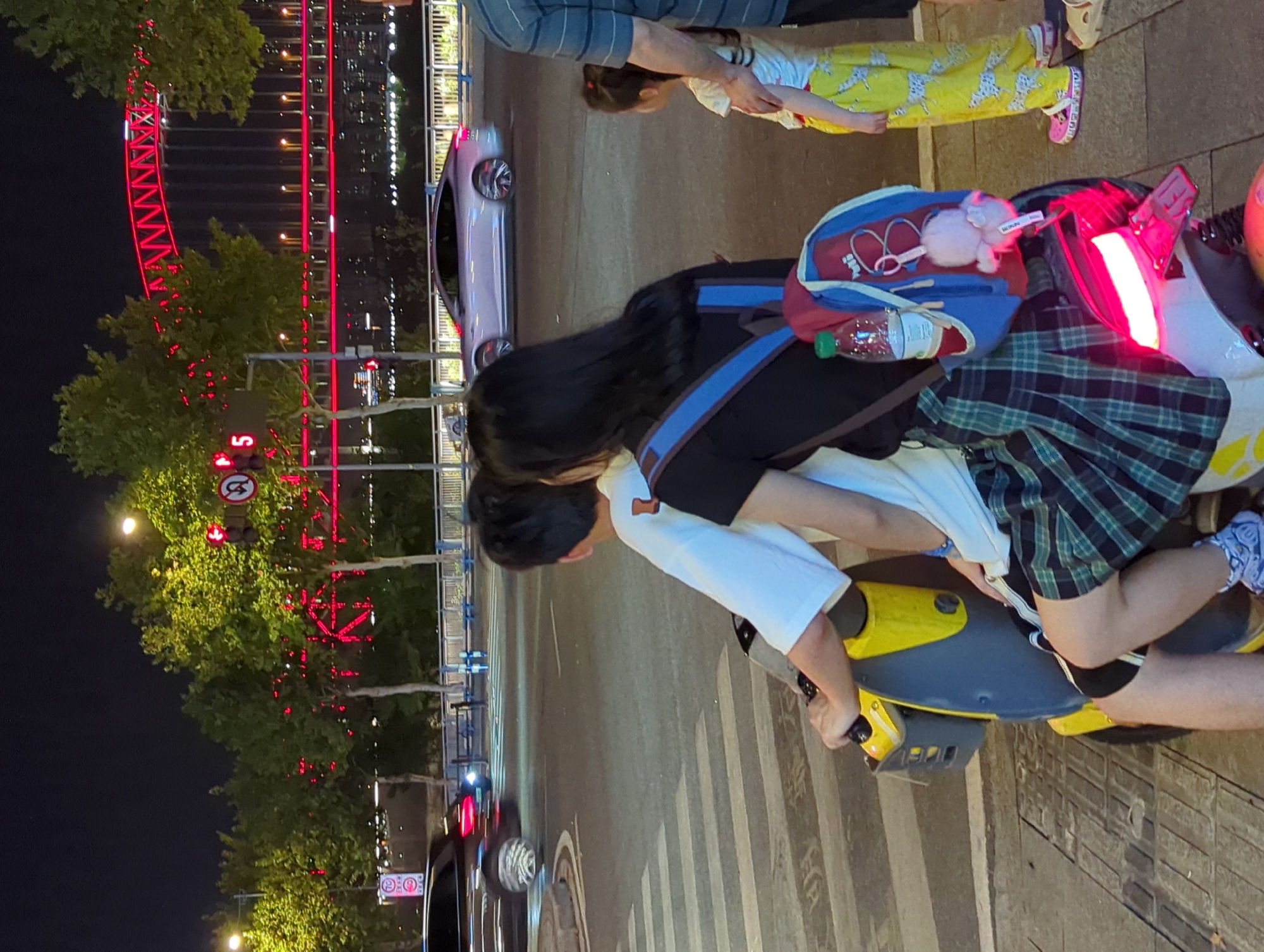 In the city of 江北城街道, China, at night, two people are riding a yellow electric scooter. The person in front, wearing a white t-shirt, is operating the scooter. The person behind them, wearing a black t-shirt and a plaid skirt, has a blue and red backpack with a water bottle and a stuffed animal attached. They are stopped at a crosswalk. In the background, a red illuminated bridge spans across a body of water. Several trees with red lights strung on them are visible. Other vehicles, including a dark-colored car and a silver sedan, are on the road. A small child in yellow patterned pants and a white shirt, accompanied by an adult, stands on the sidewalk to the right. Traffic lights are visible in the upper left quadrant of the image.