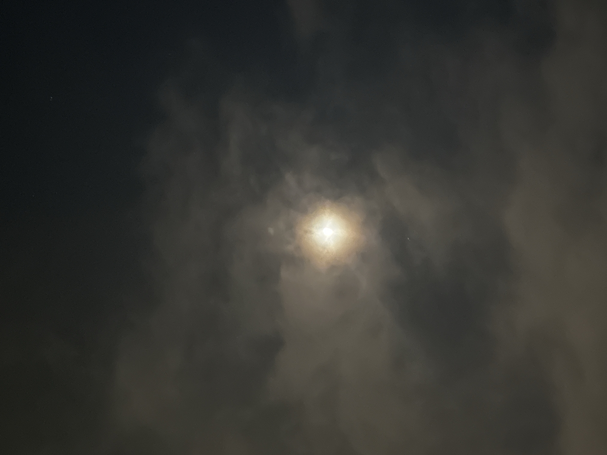 The image shows a full moon partially obscured by clouds at night. The moon is a bright, luminous orb in the center of the frame, emitting a warm, golden light that filters through the darker, more translucent clouds surrounding it. The sky is a deep, dark gray, suggesting a nighttime setting. Some faint stars are visible in the upper portion of the image, peeking through the atmospheric haze. The clouds are varied in texture and density, with some appearing wispy and others more substantial, creating a dynamic and somewhat moody atmosphere. There are no people or man-made structures visible in the image. The scene is entirely natural, focusing on the celestial phenomenon of the moon amidst a cloudy night sky. Without additional context or visual cues within the image itself, it is impossible to specifically determine the location within the city of Budva, Montenegro.