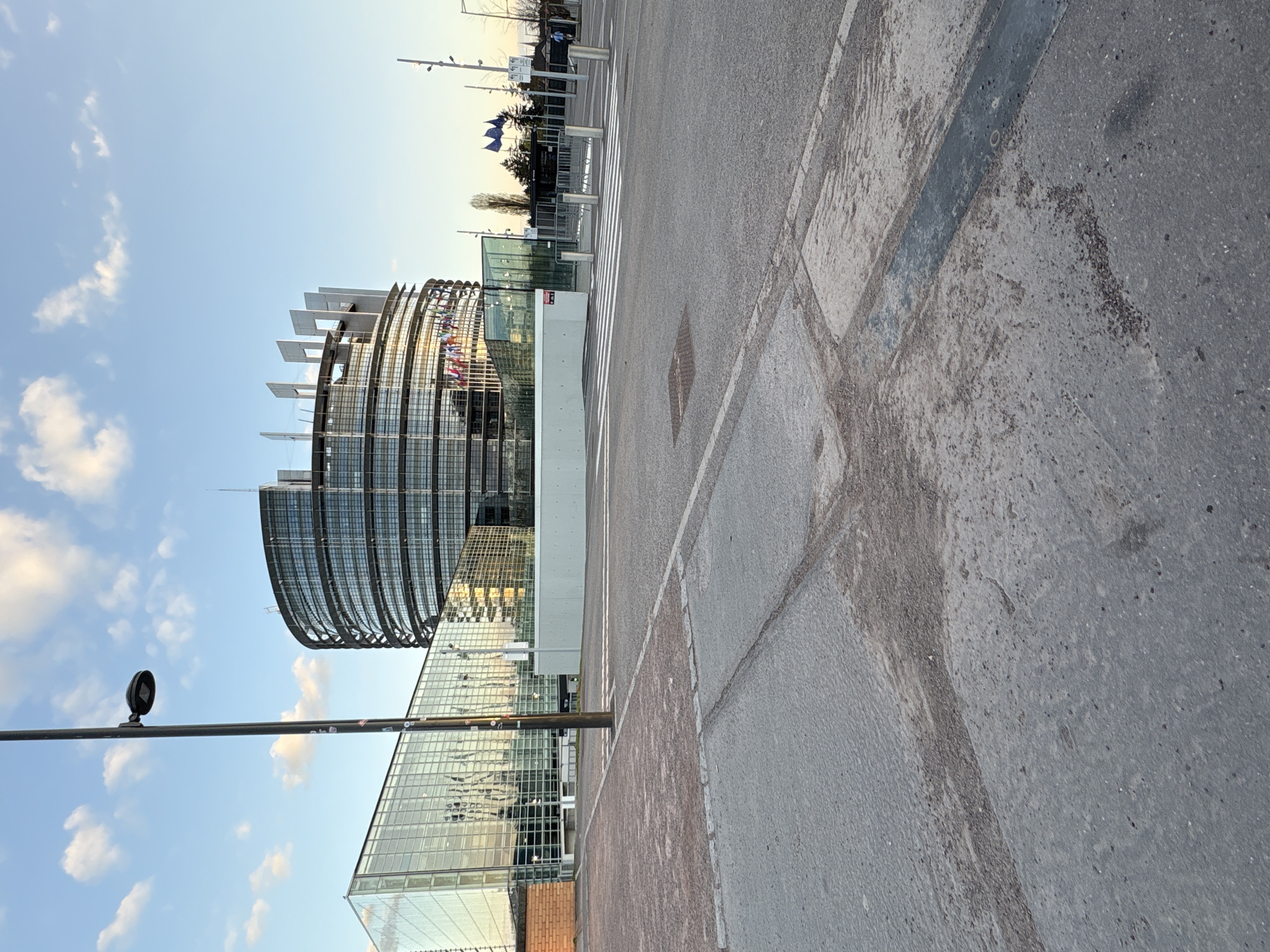 The image shows the European Parliament building in Strasbourg, France. The modern glass and steel structure is partially visible, with the main cylindrical section rising prominently against a blue sky with scattered clouds. The foreground consists of a paved area, possibly a sidewalk or plaza, with varying textures and patterns in the stone or concrete. A tall, thin lamppost stands on the left side of the frame. Further in the background, a pedestrian crossing and a row of flagpoles displaying European Union flags can be seen. The overall light suggests it is either early morning or late afternoon.