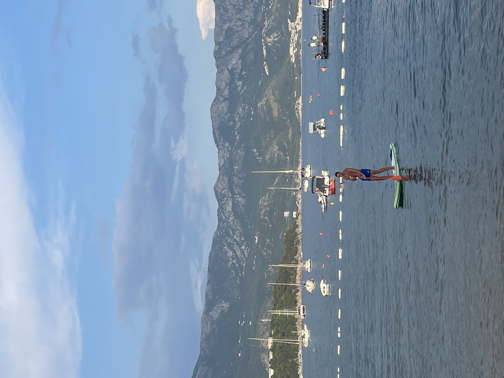 The image shows a man standing on a paddleboard in the water, with a mountain range and the town of Budva in the background. The sky is blue with some scattered clouds, suggesting it is daytime, likely late afternoon due to the lighting. The sea is calm, with several boats and buoys visible. The man appears to be enjoying himself as he paddles on the water. The scene is peaceful and serene, with the natural beauty of the mountains and the sea providing a picturesque backdrop. There is no visible text in the image.