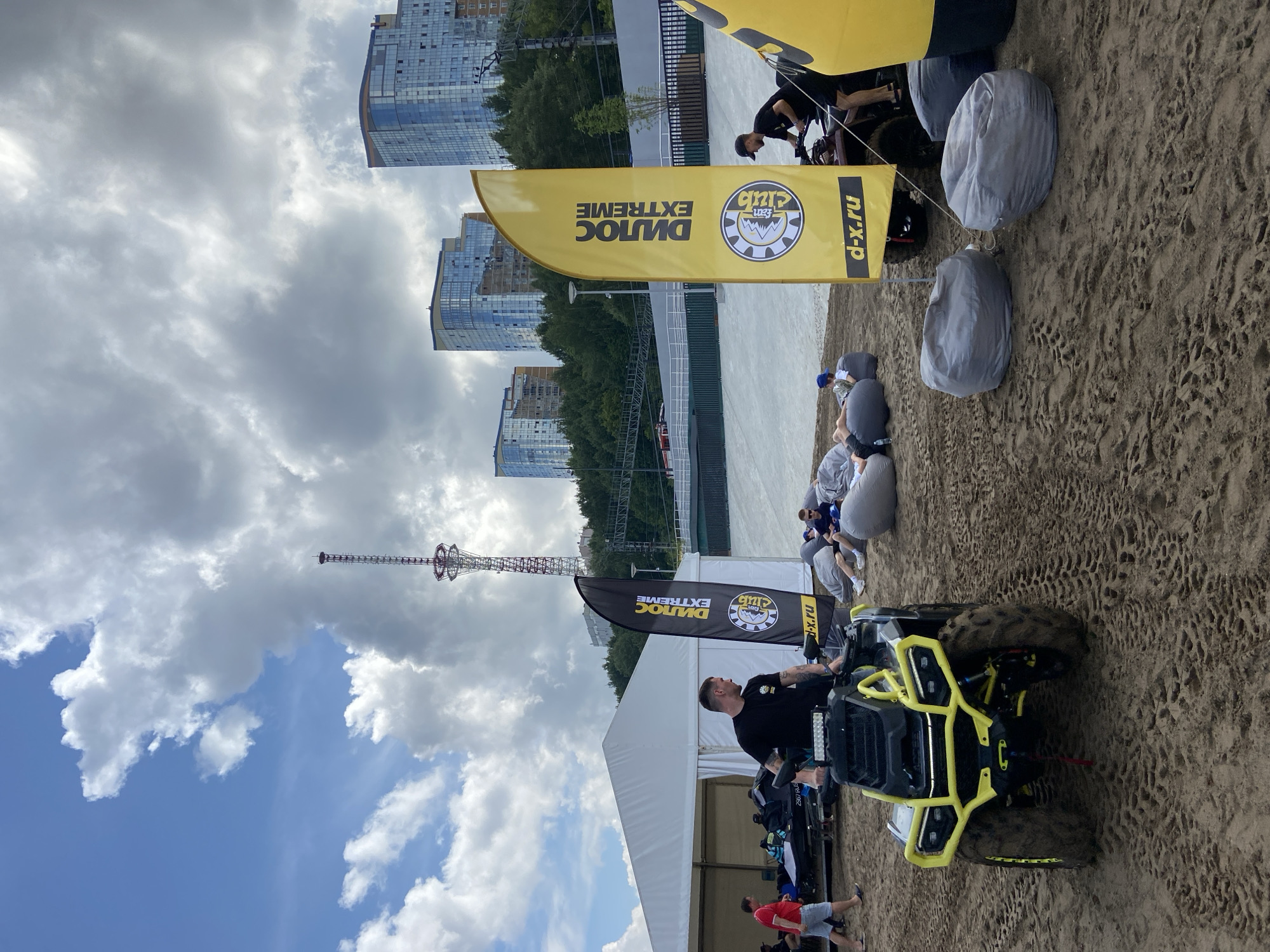 This is a medium shot of an outdoor event on a sandy area, possibly a beach, with a view of the Kama River in Perm, Russia. In the foreground, a person is standing next to a yellow and black ATV. Further back, several people are relaxing on large, grey beanbag chairs. A white tent is visible on the left, and two tall, modern buildings stand in the background, situated on a forested hillside. A large communication tower is also visible in the distance. Two yellow flags with black text and logos are prominently displayed, one of which reads "DILLOS EXTREME." The sky is partly cloudy. The overall scene suggests a promotional event or recreational gathering.