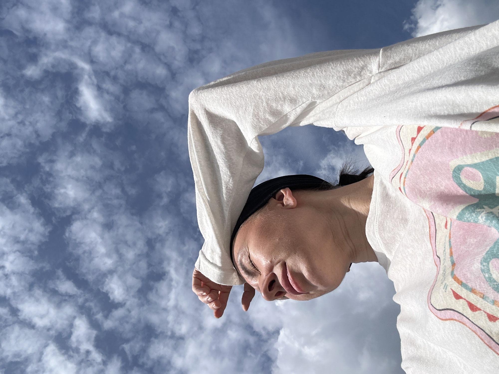 The image displays a low-angle view of a single adult female against a bright blue sky partially obscured by white cumulus clouds. The subject's head is tilted upwards, with her eyes closed or squinting, and a slight upward turn to her mouth. Her left hand is raised, with fingers slightly splayed and the palm partially covering her forehead, as if to shade her eyes from direct sunlight. She has dark hair pulled back, secured by a dark headband or hair tie. She is wearing a light-colored, long-sleeved top or sweatshirt adorned with a multi-colored graphic design on the chest area, featuring patterns in pink, orange, and light blue. The lighting suggests bright sunlight. The scene is situated in Izhevsk, Russia.