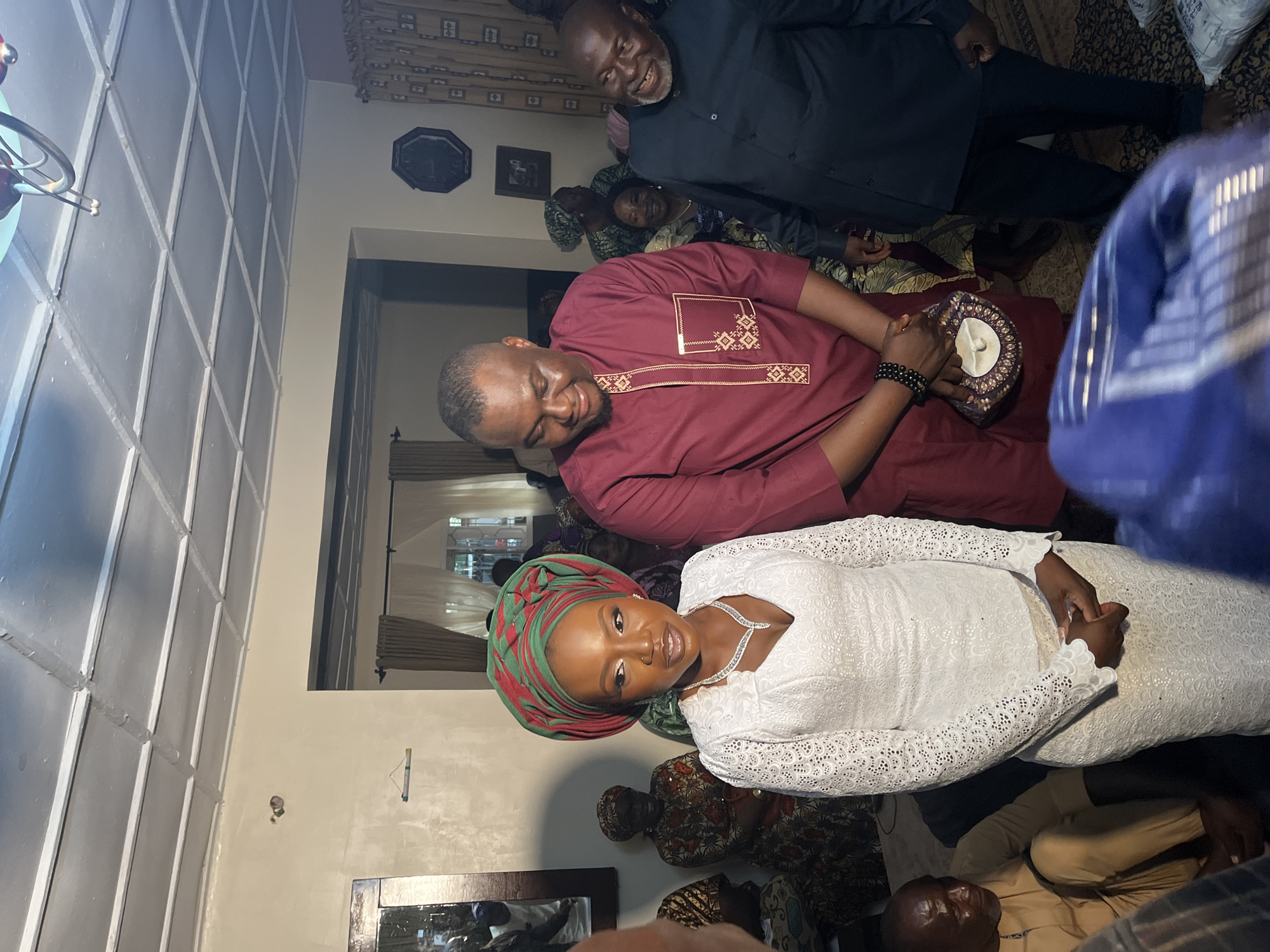 The image captures a gathering of people inside a room in Bukuru, Nigeria. The primary subjects are a woman in the foreground wearing a white lace dress and a decorative headwrap of red and green fabric, and a man standing beside her in a maroon tunic with gold trim. Both appear to be central figures, possibly at a wedding or similar ceremony.
The woman is adorned with a sparkling necklace and a nose ring. She is looking slightly to her right with a gentle smile. Her hands are clasped in front of her. The man beside her is also looking down and to his right, with a smile. He is holding a small, ornate circular clutch bag.
Behind them, other individuals are visible. To the woman's left, several women are seated, dressed in colorful traditional attire. To the right of the man in maroon, an older man in a dark blue shirt is laughing heartily, facing towards the viewer. The room has a textured ceiling with a grid pattern and a light-colored wall. Curtains are visible in the background, suggesting windows. The floor is covered with a patterned rug. The overall atmosphere appears festive and communal.