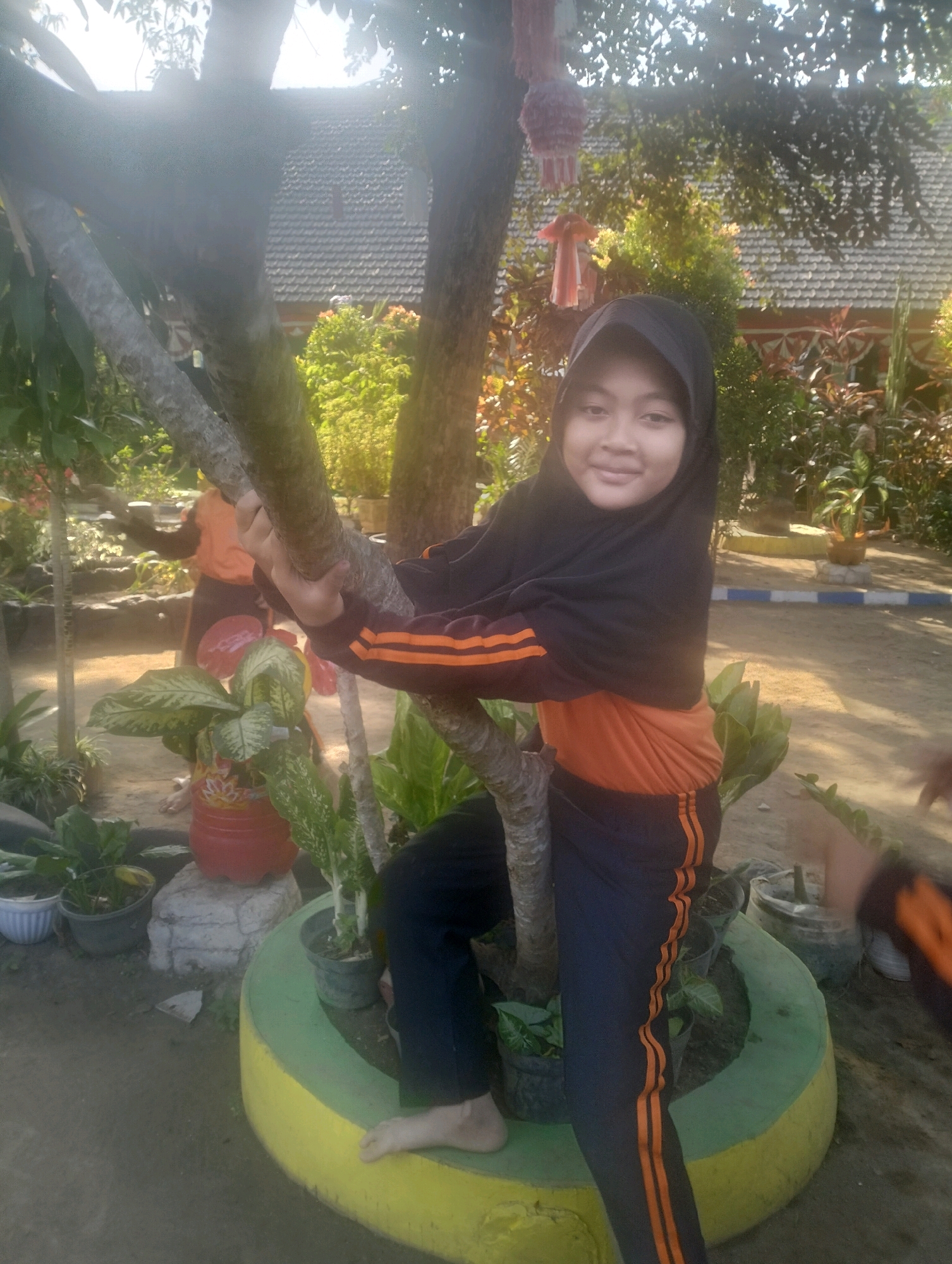 A young girl wearing a black hijab and an orange and black tracksuit stands outdoors, holding onto a tree trunk. She is positioned in the foreground, looking towards the camera with a slight smile. Behind her, another person, partially obscured, also appears to be interacting with the tree. The environment suggests a residential area with lush greenery, including potted plants and trees, and a glimpse of a tiled roof in the background. Decorative items, possibly lanterns, hang from a tree branch above. The scene is illuminated by natural sunlight, creating some lens flare. The ground is composed of dirt and pavement. The location is identified as Blabakan, Indonesia.