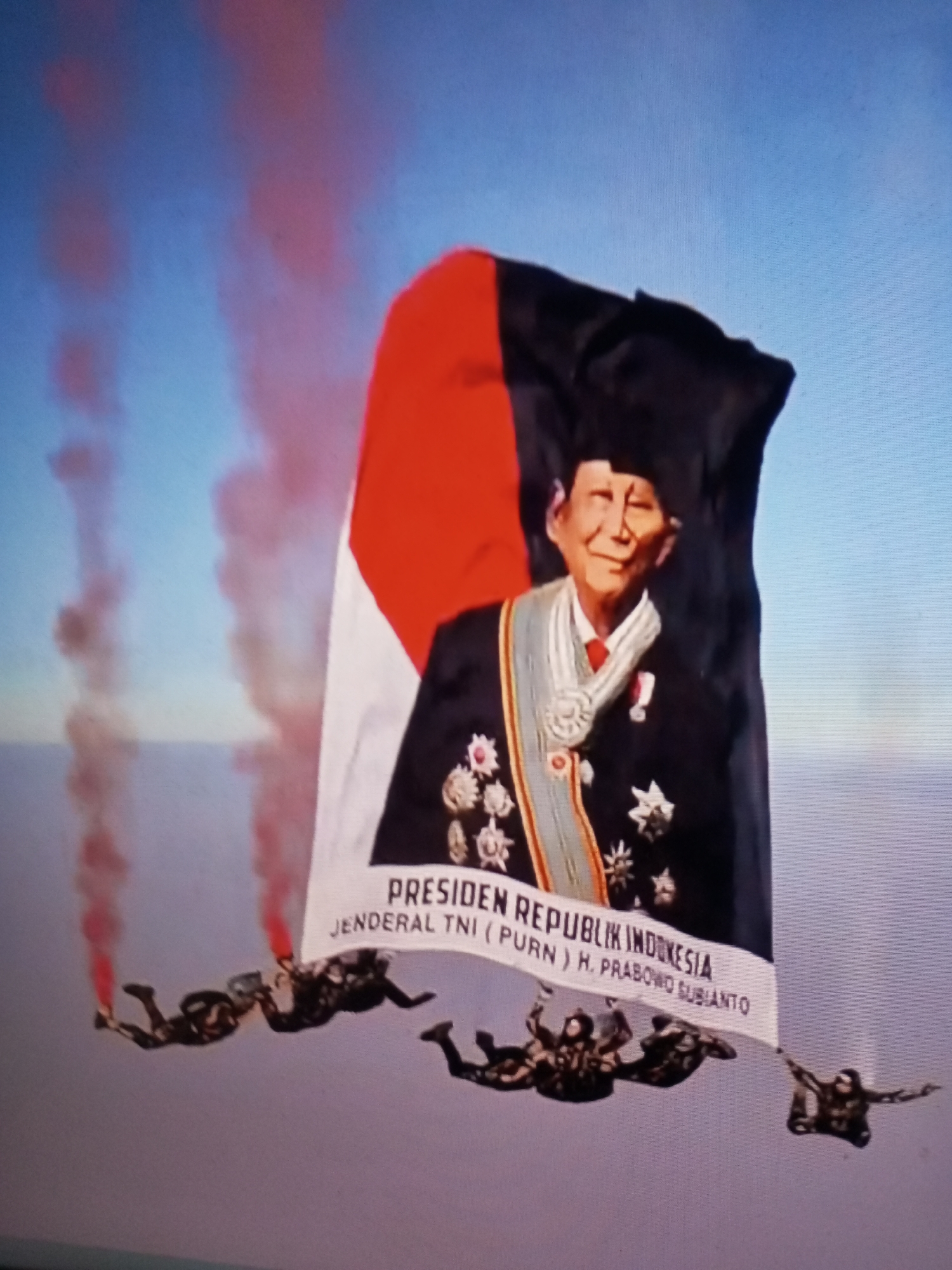 In the sky above Banyudono, Indonesia, a group of skydivers performs a stunt. They are holding onto a large banner that features a portrait of a man in military attire, along with text that reads "PRESIDEN REPUBLIK INDONESIA JENDERAL TNI (PURN) H. PRABOWO SUBIANTO". The skydivers are also deploying red smoke flares, creating a dramatic visual effect against the blue sky. The banner is partially obscured by a large Indonesian flag, with red and white sections visible. The skydivers are positioned in a formation, with some appearing to be slightly ahead of others. The overall scene suggests a patriotic or commemorative event.
