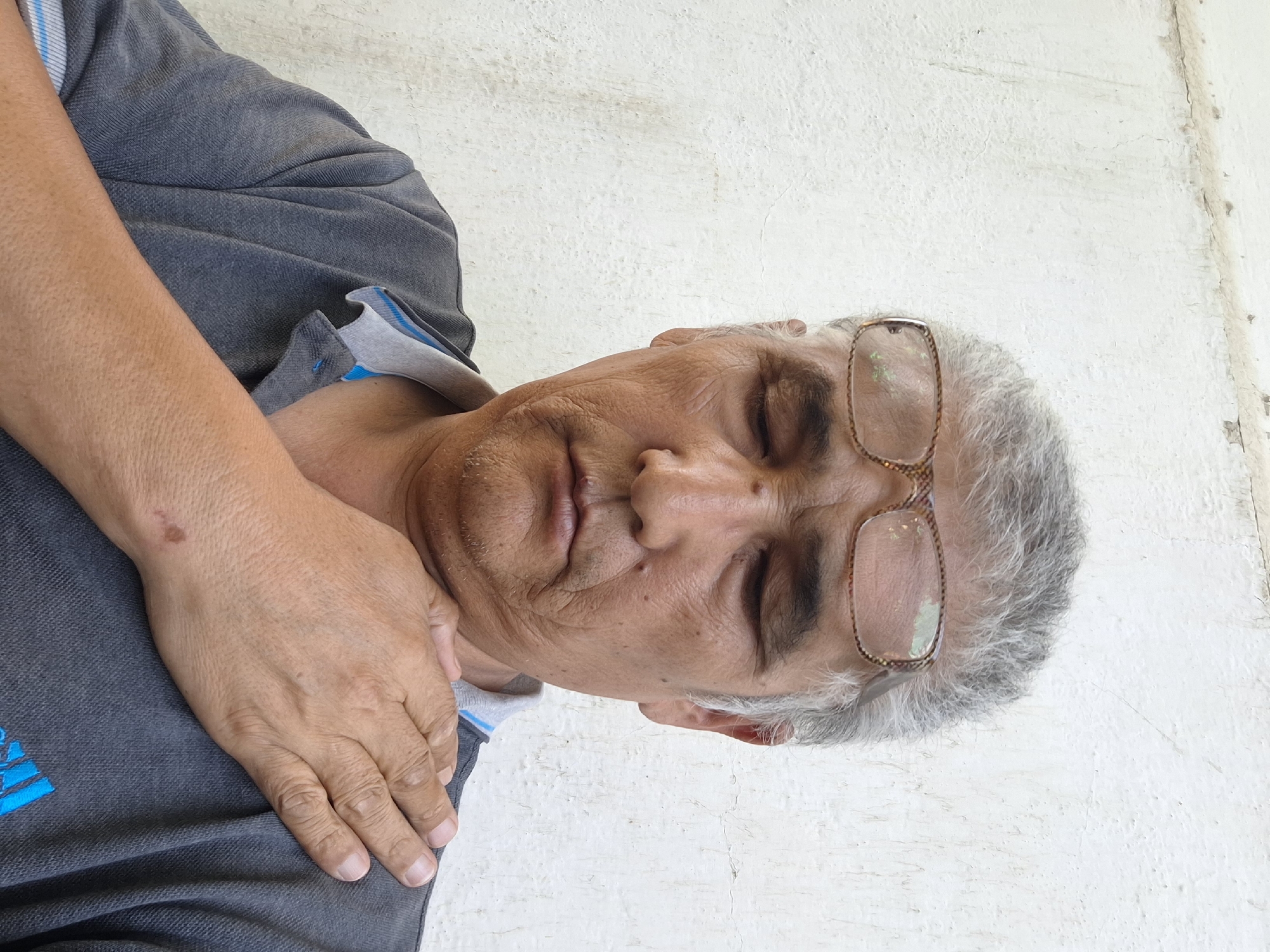 The image is a medium shot of an older man with graying hair and glasses, standing against a textured white wall. He is looking downwards with his eyes closed, and his right hand is placed on his left shoulder. He is wearing a dark gray polo shirt with blue trim around the collar. The lighting suggests an outdoor environment, possibly during the day. There are no other people or objects clearly visible in the frame besides the man and the wall. The location in Almaty, Kazakhstan cannot be determined from the visual information provided.