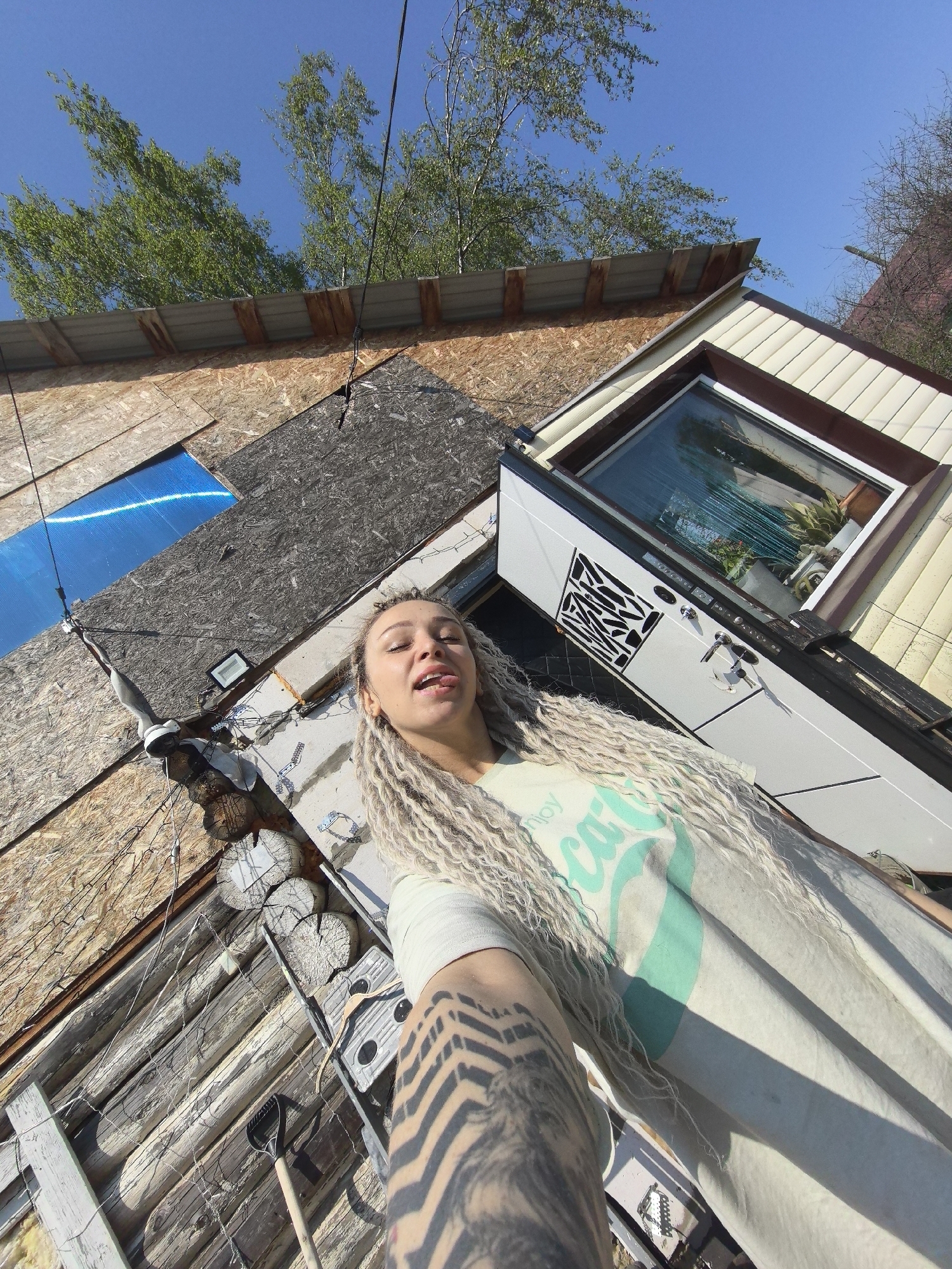 A low-angle, full-body shot presents a woman with light-colored dreadlocks sticking out her tongue, wearing a light-colored T-shirt with a turquoise graphic and intricate black zig-zag tattoos on her arm. The background features the exterior of a building constructed from a variety of materials, including wood planks, logs, plywood, and siding. A window with a dark frame and a white door with decorative cutouts are visible on the building's facade. Above, a corrugated metal roof is partially obscured by tree branches against a blue sky. Wires and cables crisscross in front of the building. The image was taken in Noviy, Russia.