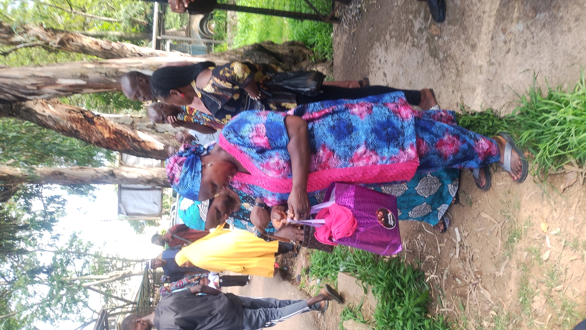 In Jos, Nigeria, a group of individuals are gathered outdoors, possibly near a street or path lined with trees. Two women in the foreground are interacting, one holding a pink shopping bag and the other reaching into a brown handbag. Both women are wearing traditional Nigerian attire. The woman on the right is dressed in a blue and pink patterned outfit with a matching headscarf. The woman to her left wears a blue and white patterned dress and headscarf. Behind them, other people are visible, including a person in a bright yellow garment and a man in a black shirt. The environment appears to be somewhat rural, with a dirt path and overgrown grass. In the background, trees with rough bark are prominent, and glimpses of structures and vehicles suggest an urban or semi-urban setting. The overall scene depicts a moment of casual gathering or activity among a community.