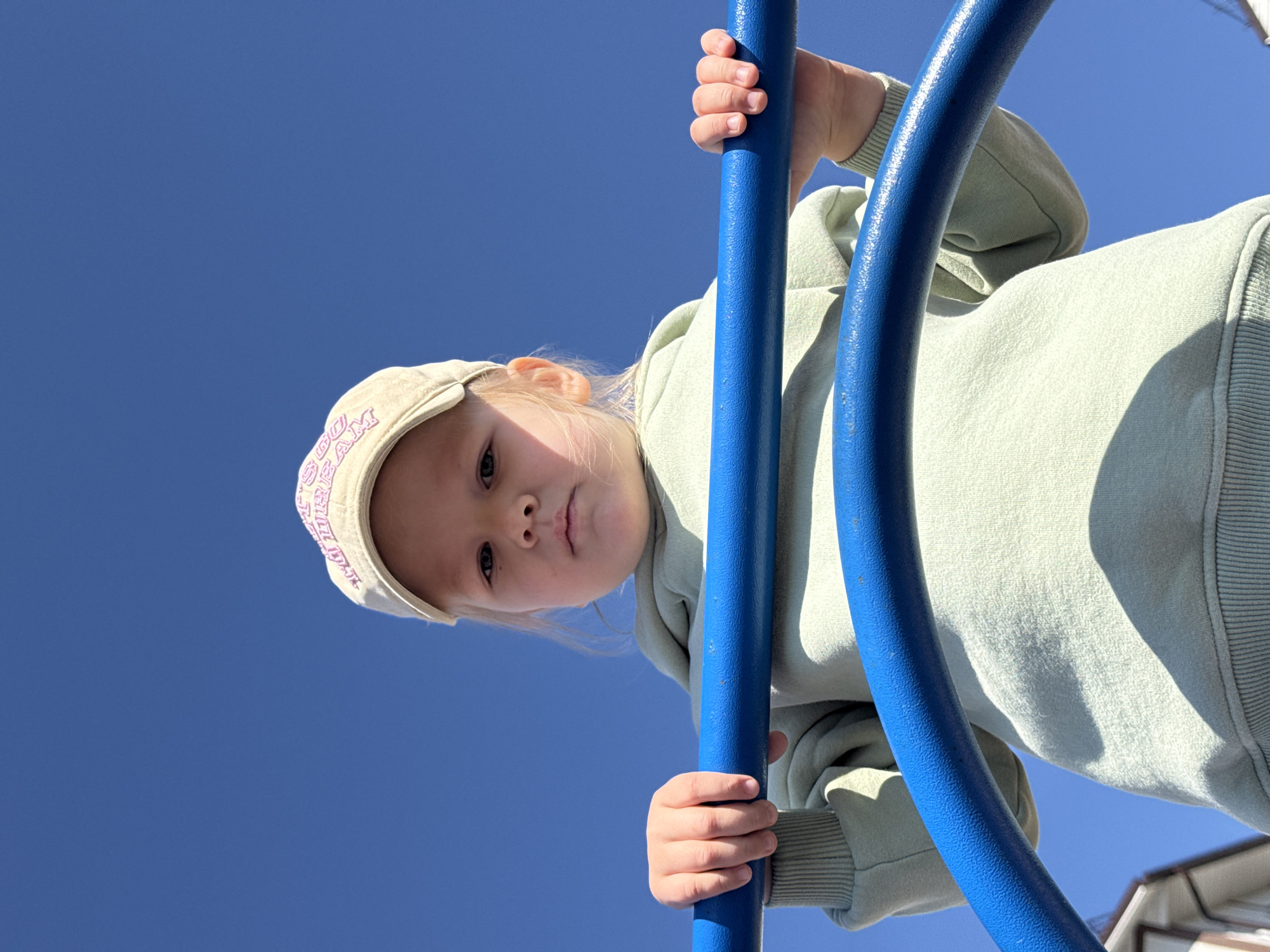 A young child wearing a light green sweatshirt and a tan baseball cap is holding onto two blue metal bars, likely part of a playground structure. The perspective is from below, looking up at the child against a clear blue sky. The child appears to be looking down at the viewer. The location is Горный, Russia. A building is visible in the bottom right corner.