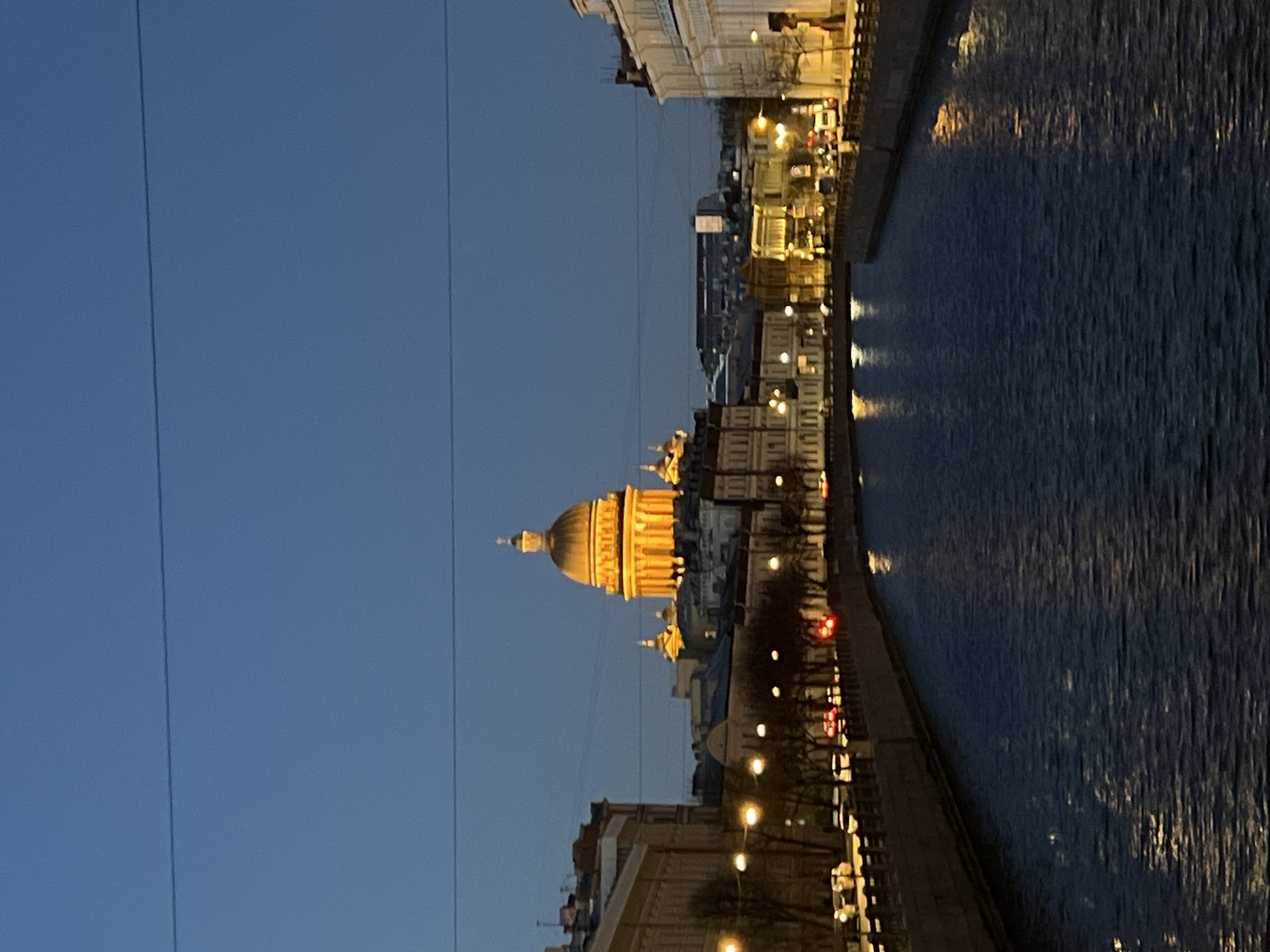 The image depicts a nighttime urban landscape in Saint Petersburg, Russia. The most prominent feature is the illuminated dome of Saint Isaac's Cathedral, standing tall amidst the surrounding buildings. A body of water, likely a canal or river, runs alongside the buildings, reflecting the city lights. The sky transitions from a deep blue at the top to a lighter hue near the horizon. Streetlights illuminate the embankment, casting reflections on the water. Power lines are visible in the sky.