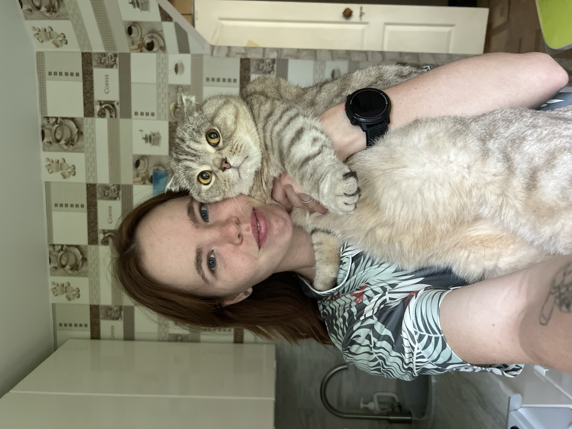 A young woman with auburn hair and blue eyes is holding a grey tabby cat in what appears to be a kitchen. She is wearing a light-colored pajama top with a tropical leaf print. The cat has large, yellow eyes and is looking directly at the camera. Behind the woman, there is kitchen cabinetry, a faucet, and a patterned wallpaper with coffee-related imagery. The scene is indoors, and the lighting suggests it is daytime. The woman is positioned in the foreground, with the cat held close to her chest. The overall impression is a casual, personal moment captured in a home environment.