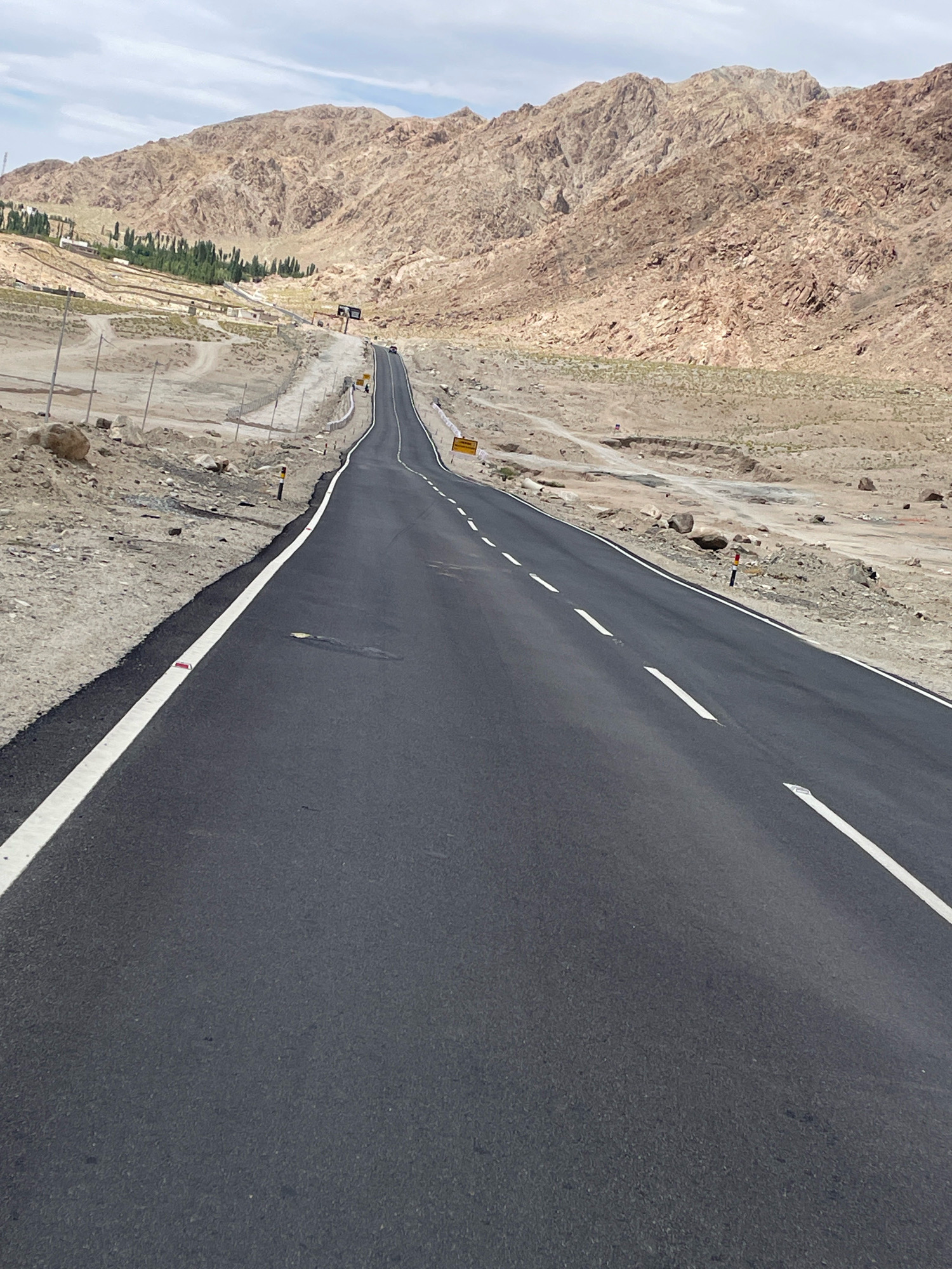 An eye-level, slightly downward-angled, vertical shot captures a two-lane asphalt road curving gently uphill, flanked by barren, rocky mountains on the right and a more vegetated hillside with buildings and trees on the left. The sky is partly cloudy. The road features white dashed lines dividing the lanes and a solid white line along the left edge, with reflective markers visible. The immediate foreground shows the dark asphalt of the road. The mountains in the background are a light brown, arid color, showing rugged, textured surfaces and sparse vegetation at their base. The left side of the road transitions to a sandy, rocky embankment with some man-made structures and a scattering of green trees. There are no people or vehicles immediately visible on the road. Yellow road signs are present in the distance. This scene is located in or around Upshi, India, a region known for its mountainous terrain and roads that traverse such landscapes.