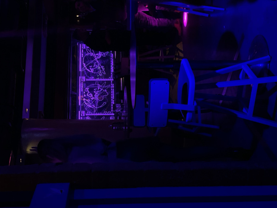 The image depicts a dimly lit, futuristic-themed indoor environment, dominated by strong artificial lighting in shades of violet and blue. The primary subjects are two vertically stacked, rectangular screens glowing with purple light, positioned on what appears to be a wall on the left side of the scene. Each screen displays a stylized world map overlaid with technological graphics, including grids, target indicators, and faint alphanumeric data. Visible text on the screens includes "TOP SECRETS", "WORLD MAP 2.0", "224 / 001 - 043", and "ELEMENTS DATA", suggesting a theme of data analysis, global intelligence, or secret operations.
To the right of the screens, several pieces of abstract or angular furniture, possibly chairs or seating, are illuminated by a bright blue light, contributing to the modern, sci-fi aesthetic of the space. A subtle pink light source is visible in the upper right background.
A person's head and shoulders are dimly visible in the lower foreground, appearing to lean over a surface or interact with the immediate environment, suggesting active engagement with the setting. Another faint reflection of a person can be seen in the upper left background.
The scene conveys the atmosphere of an immersive experience, likely an escape room, a themed exhibit, or a specialized entertainment venue. The activity taking place involves observation and interaction within this high-tech, mysterious setting. The lighting creates a sense of intrigue and technological focus. The image itself appears to be rotated 90 degrees clockwise, so the described elements would be oriented differently if viewed as a standard landscape photograph. There are no specific visual cues within the image to indicate its location in Drapetsona, Greece, other than the provided context.