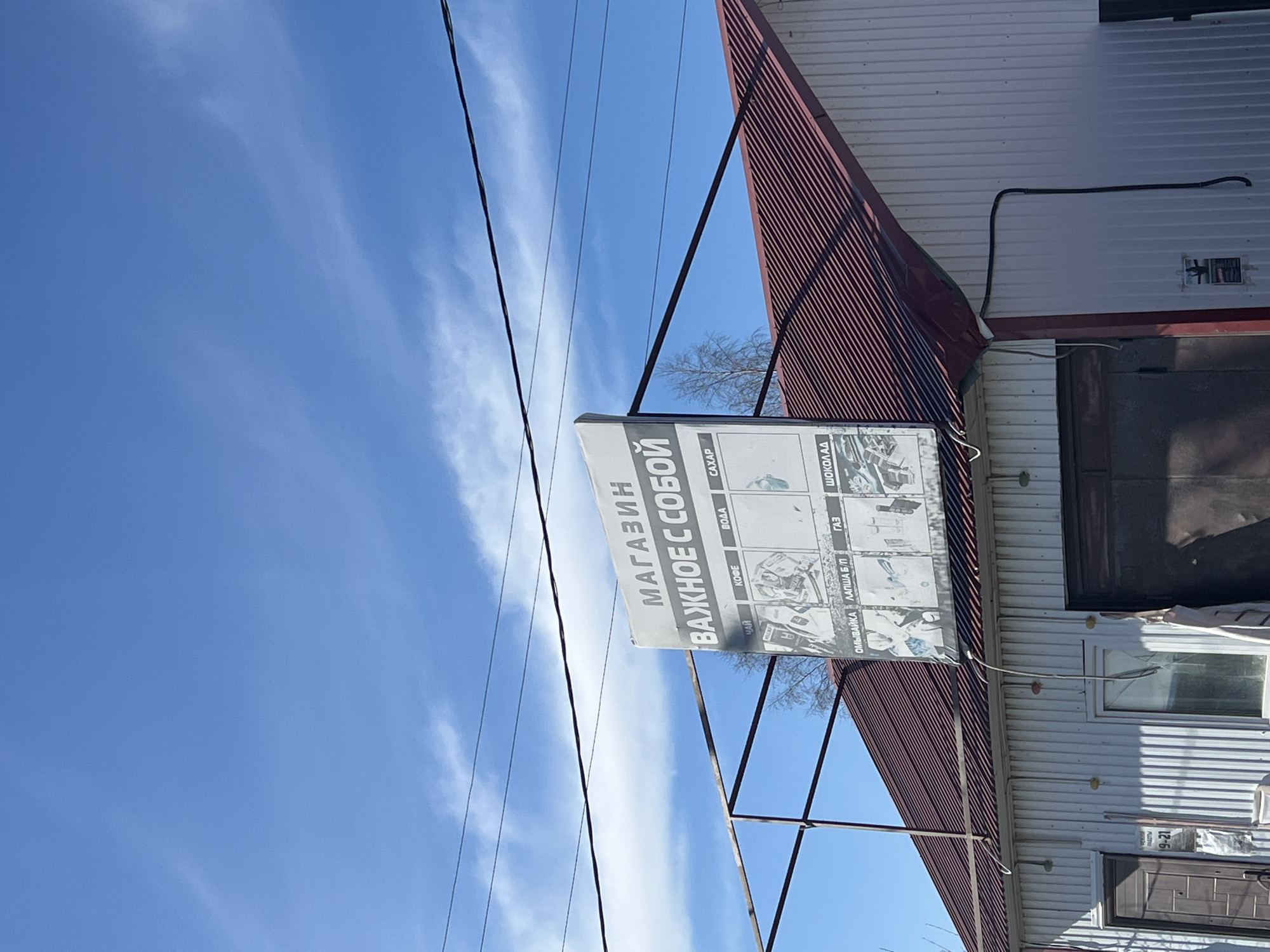 The photograph shows a building with a sign that reads "МАГАЗИН ВАЖНОЕ С СОБОЙ" (translated to "SHOP IMPORTANT WITH YOU"), with further details about items for sale below, including tea, coffee, water, sugar, windshield wiper fluid, noodles, gas, and chocolate. The building is a one-story structure with a metal roof and corrugated metal siding. There is a door and a window visible on the side of the building. Power lines run across the top left of the frame, and the sky is blue with thin clouds. The image appears to be taken in Gory, Russia, based on the language of the signage.