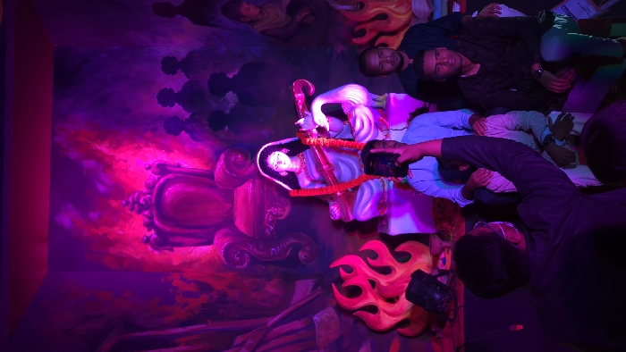 A statue of a deity sits adorned with a garland of orange flowers, next to a white swan on an ornate purple chair against a mural backdrop of pink and purple clouds. Flames sprout below. Men in casual clothing pose and interact with the statue, one holding a black object near it, while another peers behind them, some wearing masks. The lighting is dramatic, with deep purples and pinks illuminating the scene.