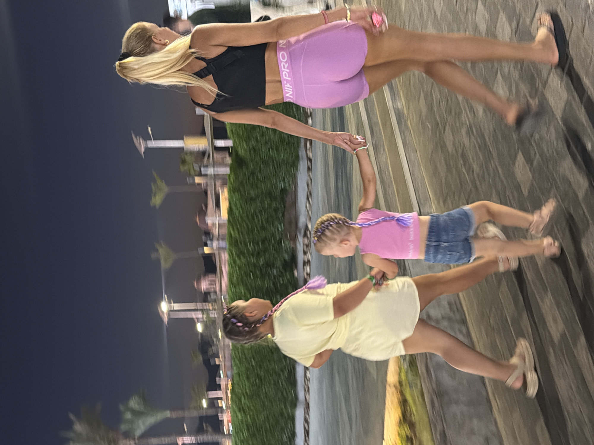 In Sharm El Sheikh, Egypt, at nighttime, three individuals, two younger girls and one woman, are walking together outdoors. The woman, with blonde hair in a ponytail, wears a black crop top and purple shorts. The older girl, to her left, is dressed in a yellow top and white shorts. The younger girl, positioned between them, is in a pink top and denim shorts. They are holding hands and walking on a paved walkway adjacent to a green hedge. Palm trees and distant lights are visible in the background, suggesting a resort or recreational area.