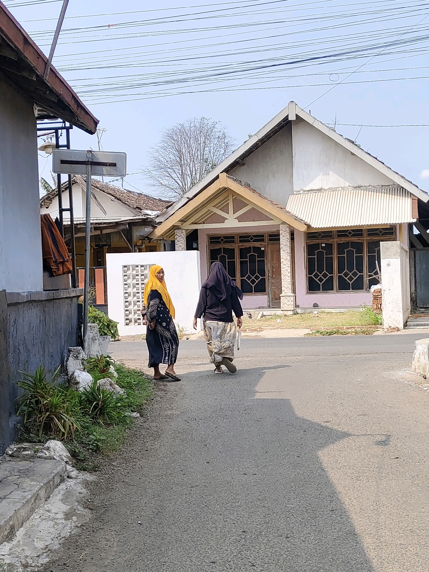 The image shows two women walking on a paved road in a residential area of Probolinggo, Indonesia. The woman on the left is wearing a yellow hijab and a dark blue patterned dress. The woman on the right is wearing a dark hijab and a light-colored patterned sarong. They are walking away from the camera towards a pink house with a white-trimmed roof and a prominent gable. The house has a large window with a decorative wooden lattice. In the background, there are other houses, some with tiled roofs, and electrical wires strung across the sky. Vegetation, including green plants and rocks, is visible along the side of the road in the foreground. The scene is illuminated by sunlight, casting shadows on the road.