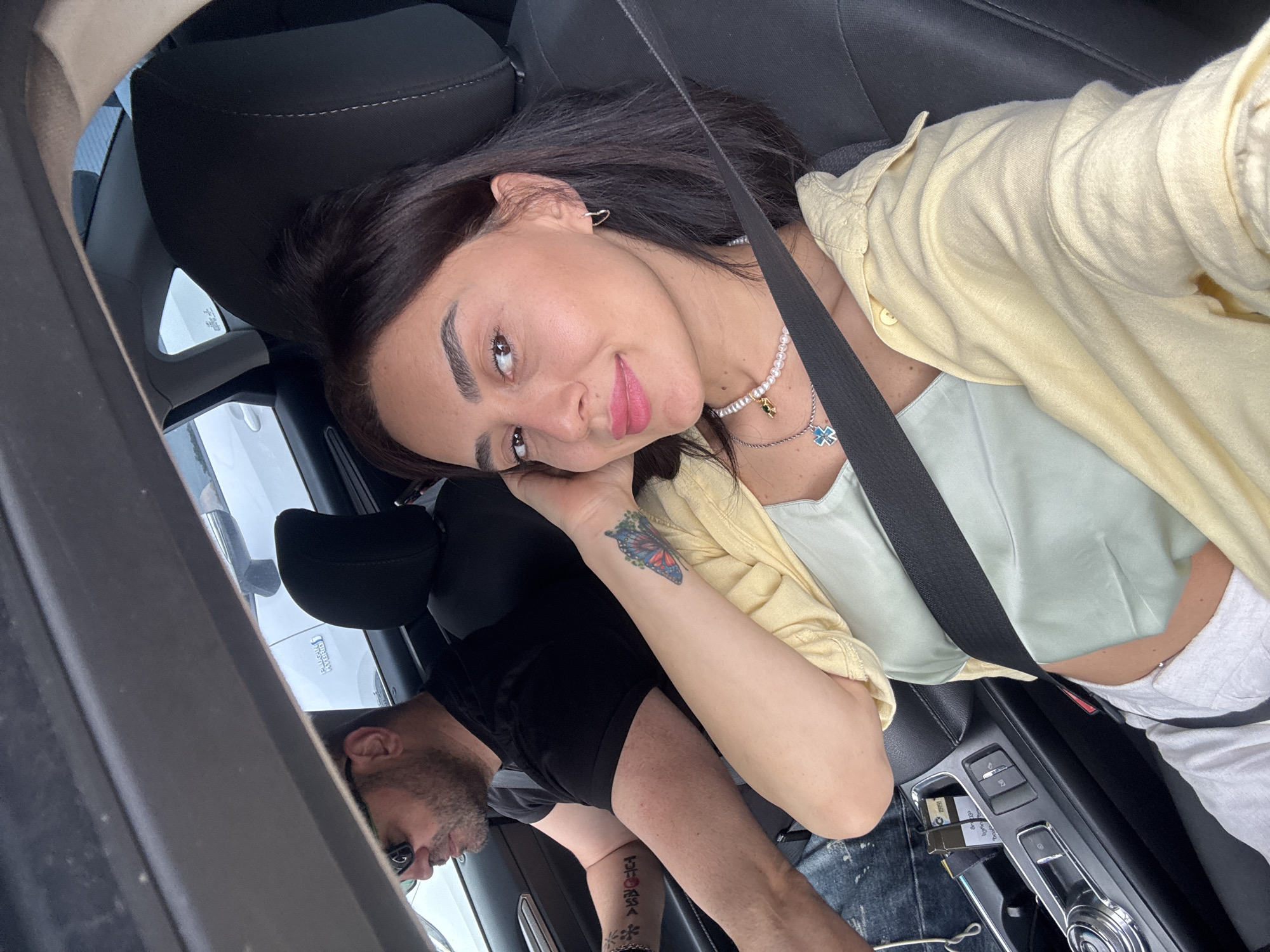 In a car, a woman with dark hair and a pale yellow open-front shirt over a light green camisole poses for a selfie. She wears a pearl necklace with a blue cross pendant and hoop earrings. Her left arm, with a butterfly tattoo, rests on her armrest. A man in sunglasses and a black t-shirt drives in the background. The car interior shows a black seat, a central console with a gear shift, and a glimpse of the exterior through the windows. The scene appears to be captured while stopped, possibly in a parking lot or traffic, given the lack of motion blur and the static nature of the background vehicles visible. The image is a close-up selfie taken from the front passenger seat. The setting is not discernable as being in Tbilisi, Georgia.
