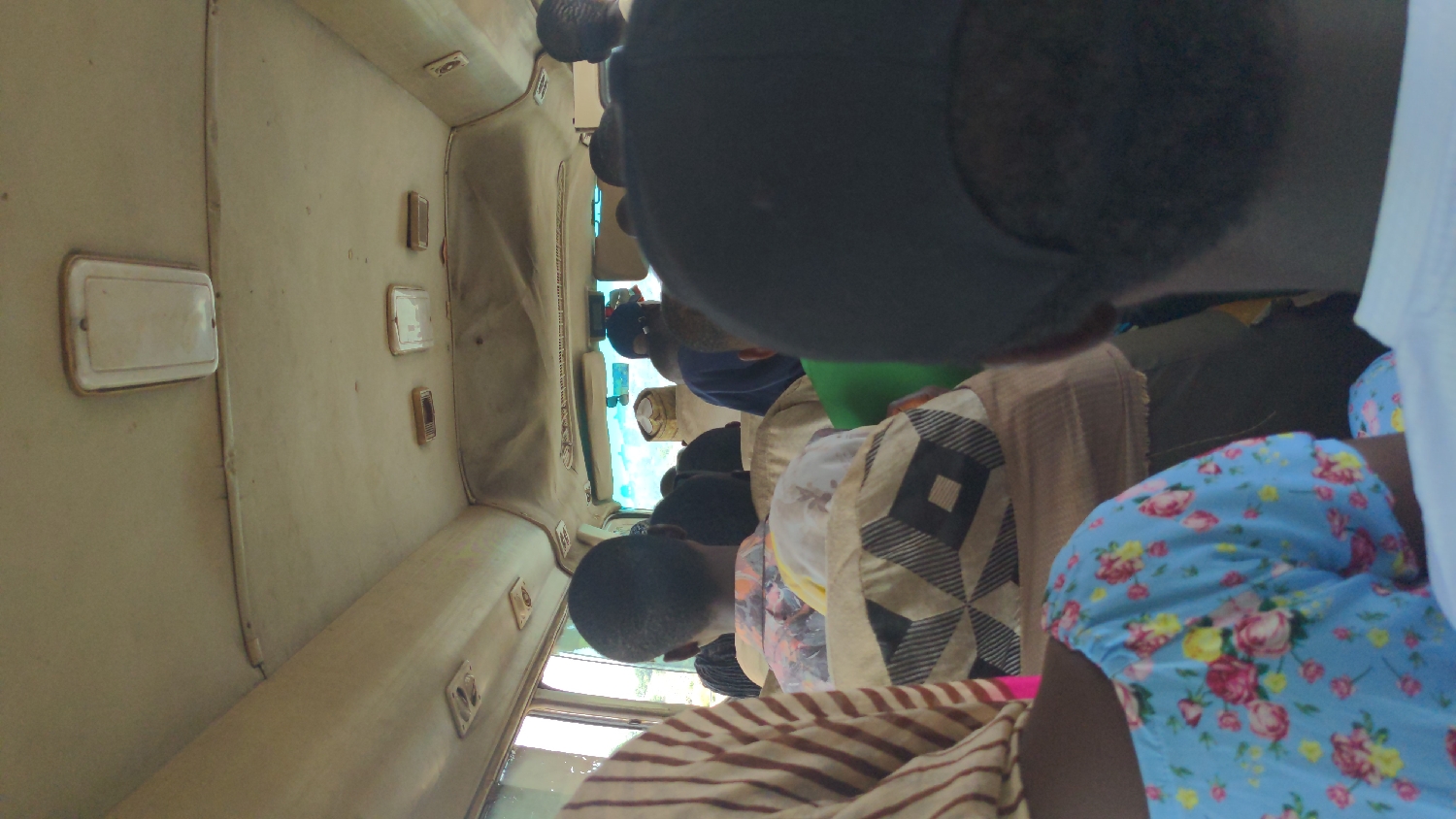 The image captures the interior of a public transport vehicle, likely a bus or large van, filled with passengers. The perspective is from behind individuals in the front seats, looking towards the front of the vehicle. The entire scene is oriented as if the camera was rotated 90 degrees clockwise.
Multiple subjects are visible, primarily from the back of their heads and shoulders, seated within the vehicle. In the immediate foreground, a person on the right wears a light blue top with a vibrant floral pattern of pink, yellow, and green flowers. Next to them, another passenger is seen wearing a garment with a distinct geometric pattern of black and off-white squares. Further ahead, several other passengers are seated, some wearing dark caps or hats. A person towards the front has a dark blue top, and a bright green garment is visible on another.
The interior of the vehicle features a light-colored, possibly off-white or cream, ceiling and upper wall lining that appears somewhat worn. Rectangular overhead fixtures, possibly lights or vents, are mounted on the ceiling. Through the front windows, a bright sky or exterior is visible, indicating it is daytime. The packed seating arrangement and vehicle type suggest a common public transportation scene in Kugbo, Nigeria. No discernible text is visible within the image. The activity depicted is simply people traveling or waiting inside the vehicle.