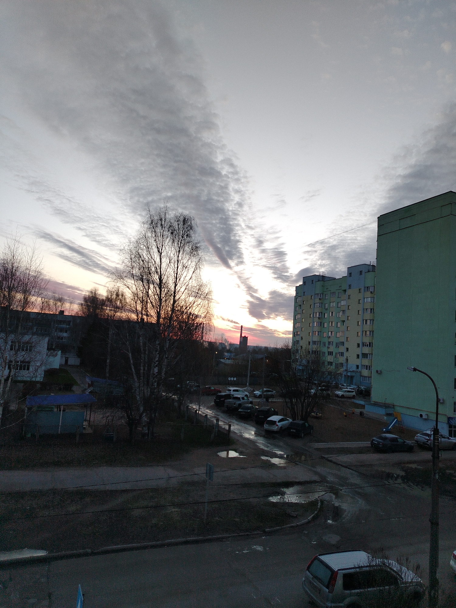 The image depicts a city scene at dusk or dawn, possibly located in an urban area of Unknown. The sky is filled with layered clouds illuminated by the setting or rising sun, casting streaks of light through the cloud cover. In the foreground, there's an intersection with visible puddles on the pavement. A silver car is parked on the road. Beyond the intersection, there is a parking area filled with several cars. Adjacent to the parking area, there's a playground area. A multi-story building with a green and yellow facade is visible on the right side of the image. Bare trees stand silhouetted against the sky, adding depth to the scene. A smaller building with a blue roof is partially visible on the left side of the image. A distant smokestack or tower is visible on the horizon. A street lamp stands on the right edge of the image. The overall impression is one of urban quietude during twilight.